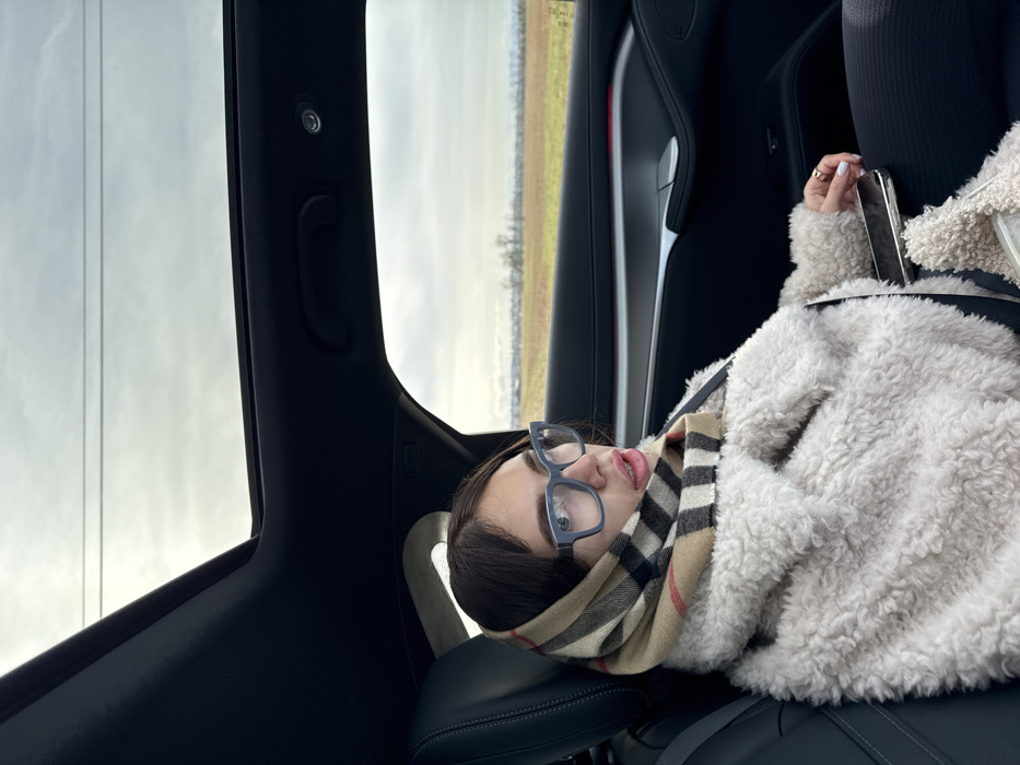 A female individual is seated in the rear passenger compartment of a vehicle, with the presumed location being Saint-Bonnet-de-Mure, France. The female, with dark hair partially covered, wears light blue-framed eyeglasses and a beige, black, and red plaid pattern scarf draped over her head and neck. She is dressed in an off-white, textured, possibly faux fur, jacket over dark trousers. A seatbelt is secured across her chest and lap. A gold-colored ring is visible on her right ring finger, and her fingernails are painted a light color. A black smartphone rests on her lap. The vehicle's interior features dark upholstery, a panoramic sunroof, and a dark door panel with a silver-colored trim and a faint red accent light. Outside the window, an overcast sky with power lines is visible above a flat landscape consisting of brown agricultural fields and distant bare trees. The individual is looking towards the left side of the frame with her mouth slightly parted.