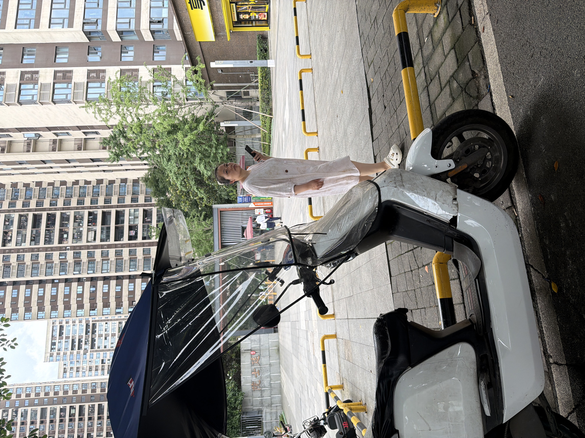 A woman stands on a sidewalk next to a white electric scooter with a black and clear plastic windshield, in the Chongqing High-tech Zone, China. She is wearing a light-colored dress and white shoes and holds a mobile phone in her left hand. The sidewalk is paved with gray tiles and bordered by yellow and black bollards. In the background are tall apartment buildings.
