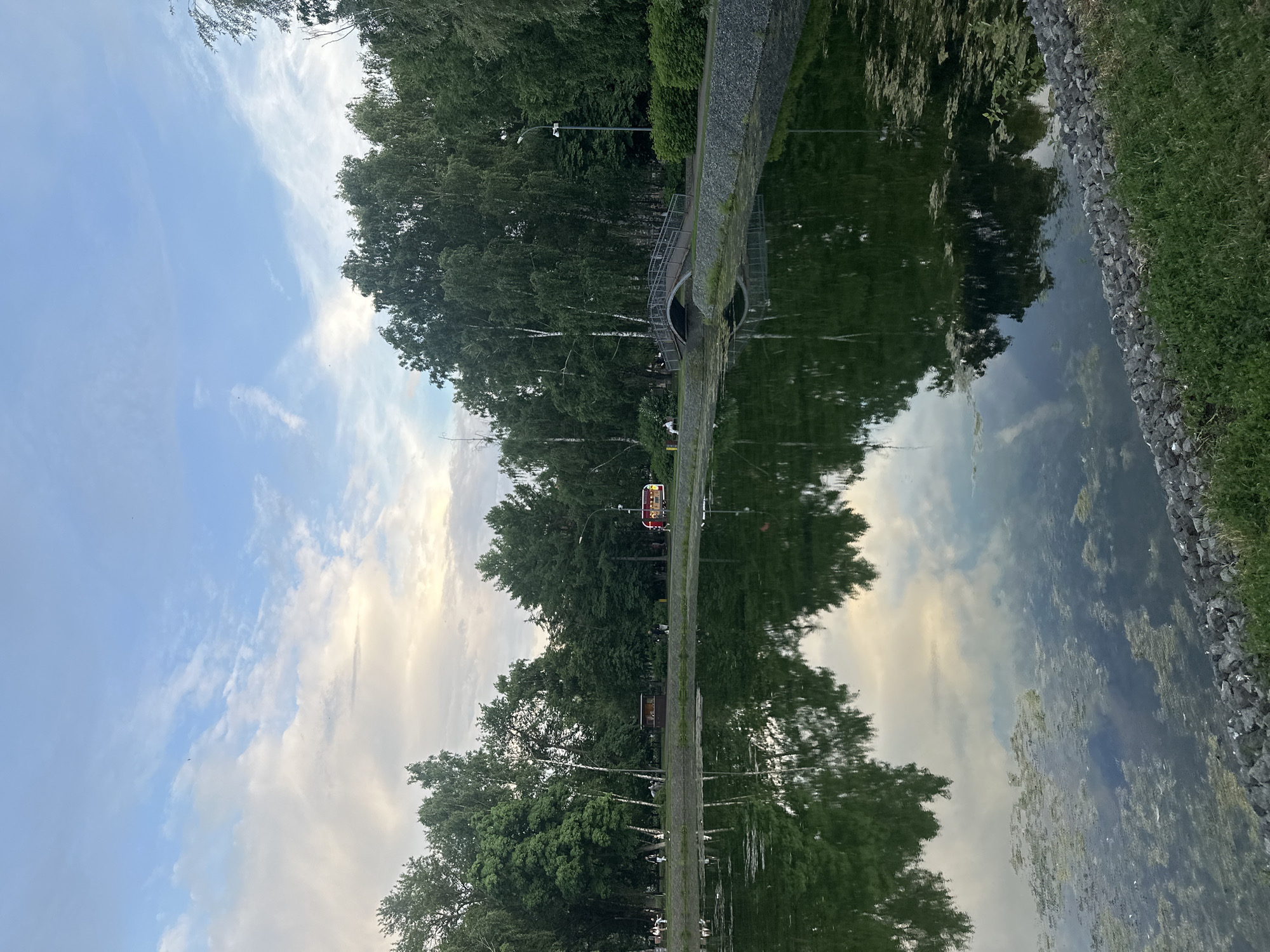 The image depicts a tranquil park scene with a reflective pond in the foreground. The pond's dark green water mirrors the surrounding trees and the sky, which is partly cloudy with soft hues of blue and white. A paved path runs along the edge of the pond, bordered by rocks and lush green grass. Beyond the path, a line of various trees, including birch trees with their distinctive white bark, creates a dense green backdrop. In the distance, a small arched bridge spans a portion of the pond, and several people can be seen strolling along the path and sitting on benches. A red and yellow kiosk or small vehicle is also visible near the path. The overall atmosphere is serene and natural, suggesting a public park setting within a city, likely Kazan, Russia, given the context. The scene is captured at a time when the light is soft, possibly late afternoon or early evening, with the sun's glow subtly illuminating the clouds.