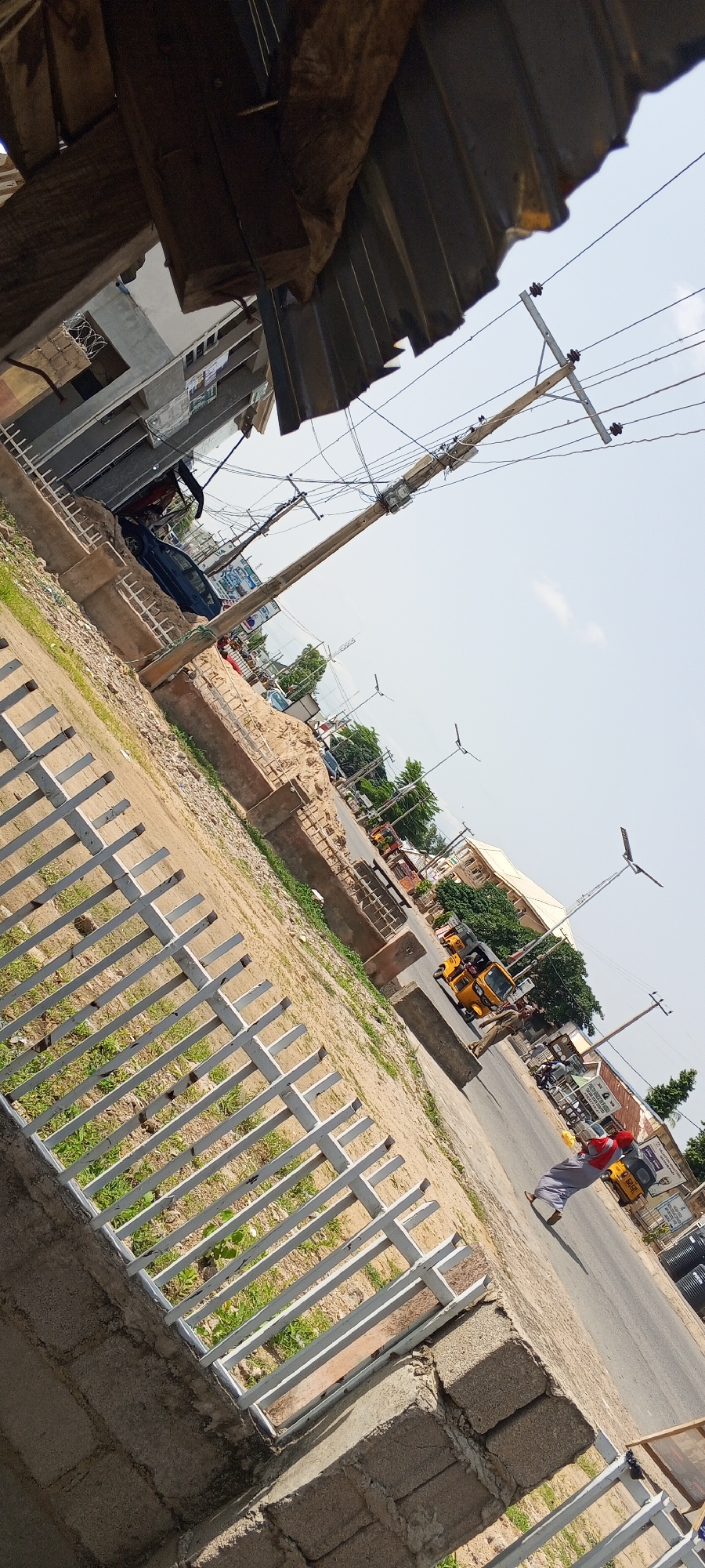 The image shows a street scene in Jalingo, Nigeria, tilted at an angle. In the foreground, a white metal fence runs along a concrete wall. Beyond the fence, a dirt and gravel area is visible, with some patchy green grass. A dark-colored car is parked on the side of the road, partially obscured by a concrete structure. Further down the road, several yellow auto-rickshaws, known as "Keke Napep," are present, some with passengers. A person wearing a red headscarf and grey clothing is walking on the side of the road, near one of the auto-rickshaws. Utility poles with numerous wires crisscross the sky above the street. In the background, there are buildings, trees, and more utility poles. The sky is clear and bright, suggesting a sunny day. The overall impression is of a busy urban street in Nigeria.