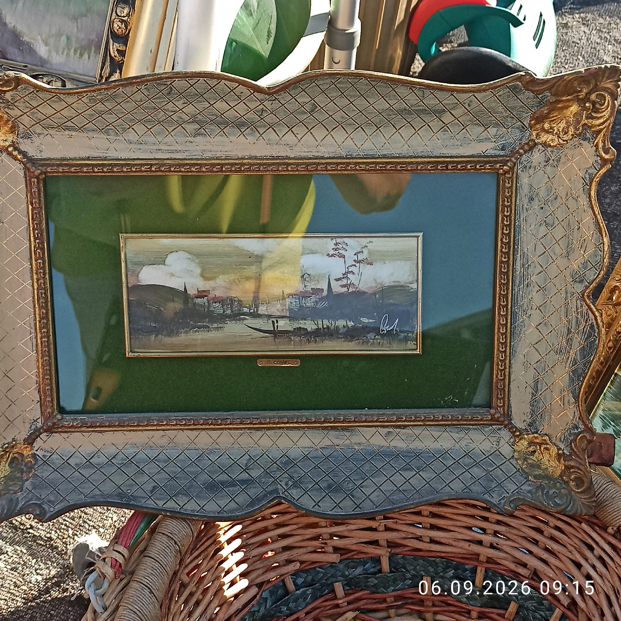 The image shows a framed painting displayed outdoors, possibly at a flea market or street stall. The painting itself is a small, horizontal landscape depicting a body of water with a boat and a village or town on the shore. The frame is ornate with gold accents and a patterned surface. In the background, there are reflections and other objects, including parts of other frames and what appears to be colorful plastic items. The bottom of the image shows a wicker basket and a timestamp "06.09.2026 09:15". The location is Sannicandro di Bari, Italy. There are no people visible in the image.
