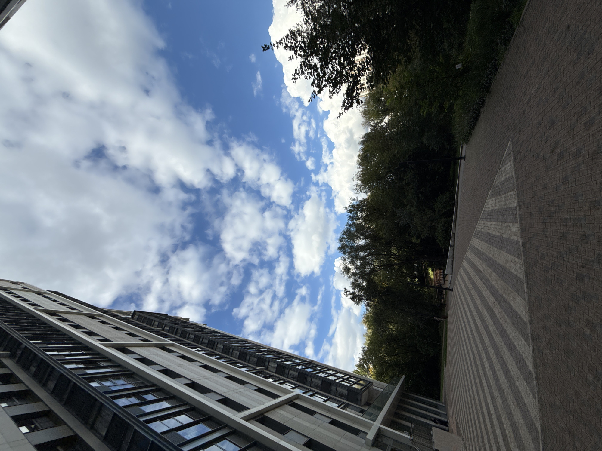 This is a ground-level, eye-level shot of a modern, tall residential building in Moscow, Russia, captured on a partly cloudy day. The building, which takes up the left side of the frame, is characterized by its sleek design, featuring numerous windows with dark frames and light-colored facade panels. The perspective is tilted upwards, emphasizing the height of the structure. In front of the building, a paved area displays a striking geometric pattern of alternating light and dark gray rectangular tiles, creating a checkered effect that leads towards a tree-lined park. The park, filled with lush green trees, occupies the background and extends across the right side of the image, contrasting with the urban architecture. The sky above is a vibrant blue, dotted with fluffy white clouds, indicating fair weather. A lamppost is visible near the edge of the park. The overall impression is one of contemporary urban development integrated with green spaces.