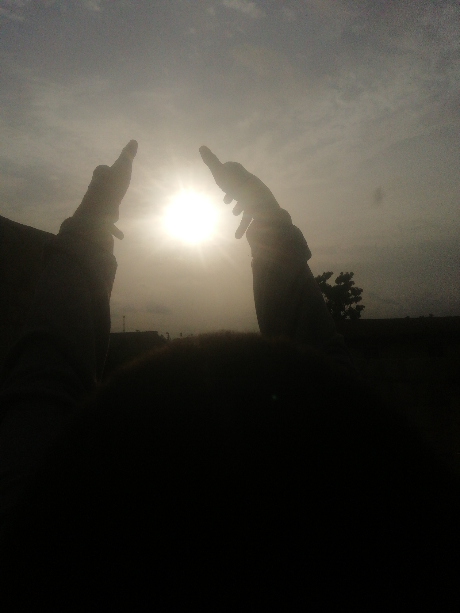 The image shows two arms raised in silhouette against a bright sun in a cloudy sky. The landscape in the background is dark, with a few trees visible. The foreground is mostly obscured by darkness. It is impossible to know the specific location in the city of Unknown, Unknown.