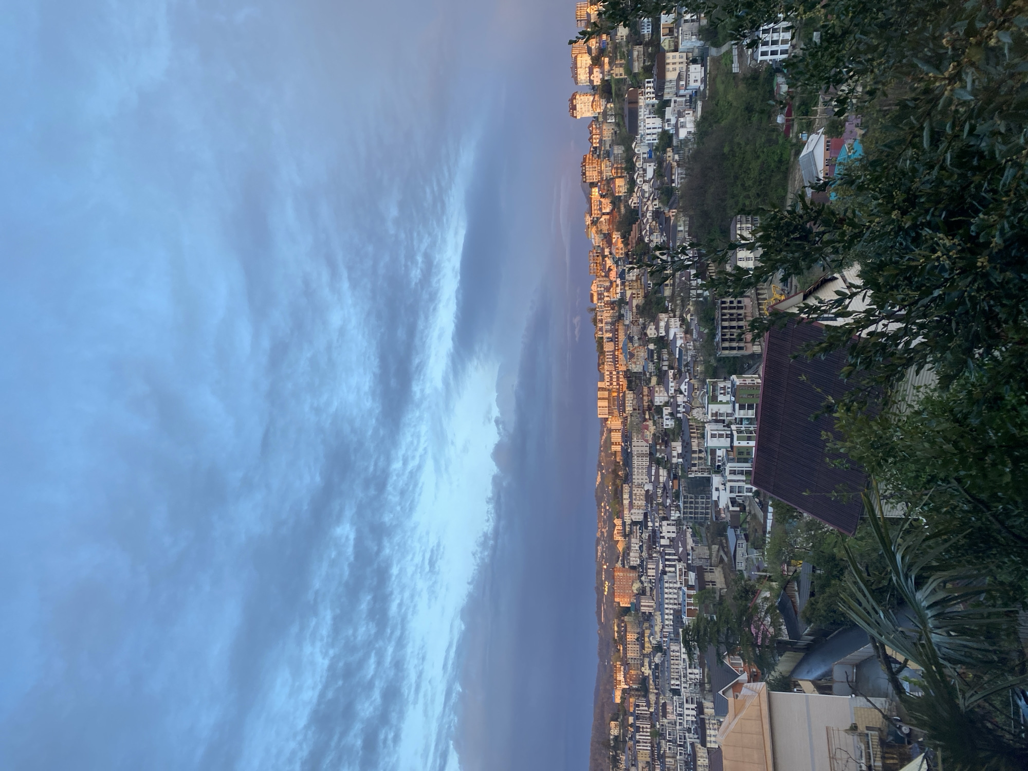 An aerial view looking up shows the city of Sochi, Russia during the golden hour. The sky features wispy clouds on the left side of the image. A dense urban area with buildings in a vertical line running up the middle, and vegetation is visible on the right.