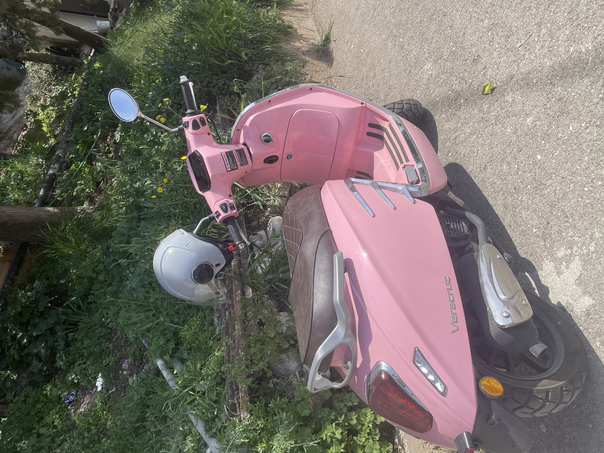A pink "Veracruz" scooter is parked on a paved surface next to a grassy area with scattered vegetation and some small yellow flowers. A gray helmet rests on the ground near the scooter's handlebars. The location appears to be outdoors in a residential area with some tree coverage in the background. No people are visible. The image could be taken in the city of Budva, Montenegro.