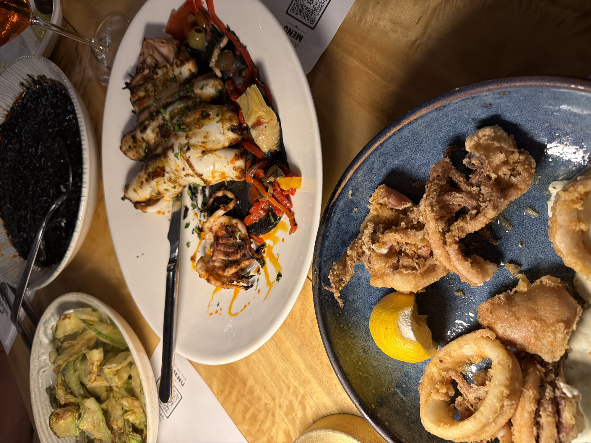 This overhead shot presents a diverse array of Mediterranean dishes laid out on a wooden table, likely in a restaurant setting in Makri, Greece. The scene is dominated by several plates of food.
In the center, a large white oval plate features grilled calamari or squid, seasoned and garnished with roasted red and yellow bell peppers, green olives, and artichoke hearts, all drizzled with an orange-yellow sauce and fresh herbs. A silver knife rests on this plate.
To the right, a rustic blue ceramic plate holds a generous serving of crispy fried calamari, including both rings and tentacles. A bright yellow lemon wedge and a portion of white dipping sauce (likely aioli or tartar) complete this dish.
On the left, a white oval bowl contains a dark, rich dish, possibly a squid ink risotto or black rice, served with a metal utensil. Below this, another white oval bowl offers a pile of lightly fried, green vegetable slices, resembling zucchini crisps.
In the upper left corner, a glass filled with a reddish beverage, possibly rosé wine, is visible. The table is made of polished light wood. Several white paper menus, featuring QR codes and the text "SCAN FOR MENU," are partially visible underneath the plates, confirming the restaurant environment. The overall impression is of a plentiful and inviting seafood meal.