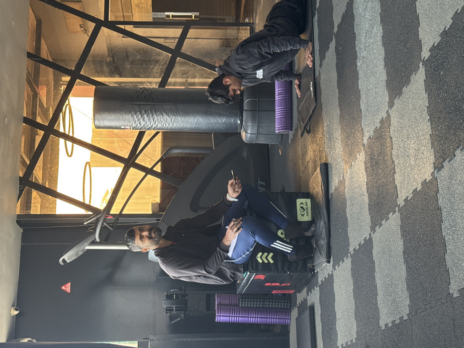 Inside a gym with black and gray checkered flooring, two people are present. One person in a dark hooded top is doing push-ups on a black mat. The other person, also in dark clothing, is seated on a black box, using a phone. A purple foam roller sits near the seated person. A piece of gym equipment, possibly a rowing machine, is in the background. Large windows let in light, showing a glimpse of the outside.