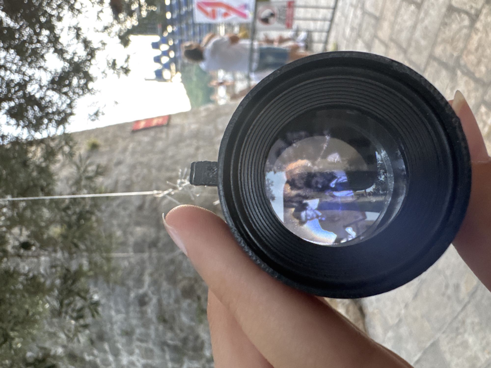 A person's hand holds a camera lens up to the viewer. Reflected in the lens are two figures, one wearing a hat and suit, and the other wearing a dark garment. In the background, a stone tower and trees are visible. Beyond that, blurred figures and structures suggest an outdoor event, possibly a festival, with stage lighting in the distance. The scene is set in the historic Old Town of Budva, Montenegro, characterized by its ancient stone architecture and bustling atmosphere.