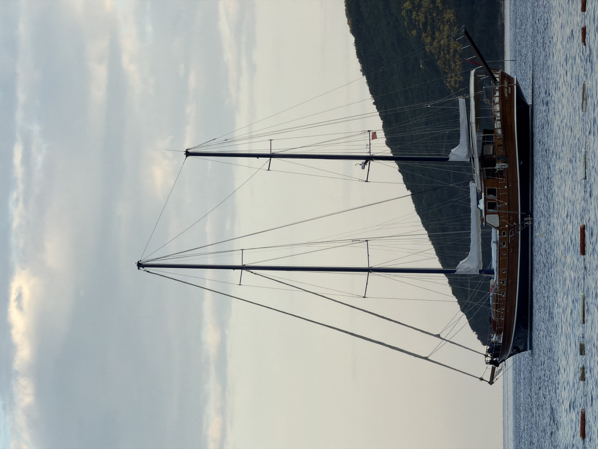 The image depicts a two-masted sailing yacht at anchor. The yacht has dark blue topsides and varnished wooden deck structures. Its sails are furled. The masts are tall and black. The backdrop is a hillside covered in green vegetation. The water is calm. The scene is possibly in the vicinity of Budva, Montenegro, based on the vegetation on the hillside, the sea, and the sailing yacht, which are typical of the region.