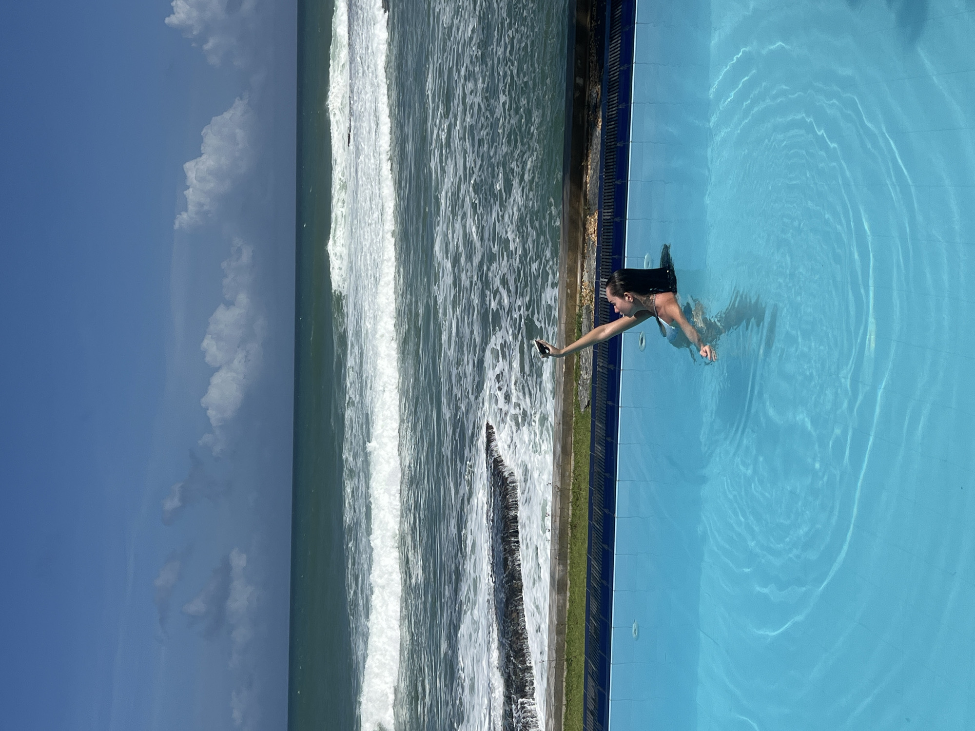 The image shows a woman in a swimming pool, holding a smartphone to take a selfie. The pool is blue and tiled, with a ledge and some greenery beyond. In the background, there is a rocky coastline with waves crashing onto it, and the ocean stretches to the horizon under a partly cloudy blue sky. The location is identified as Goviyapana, Sri Lanka.