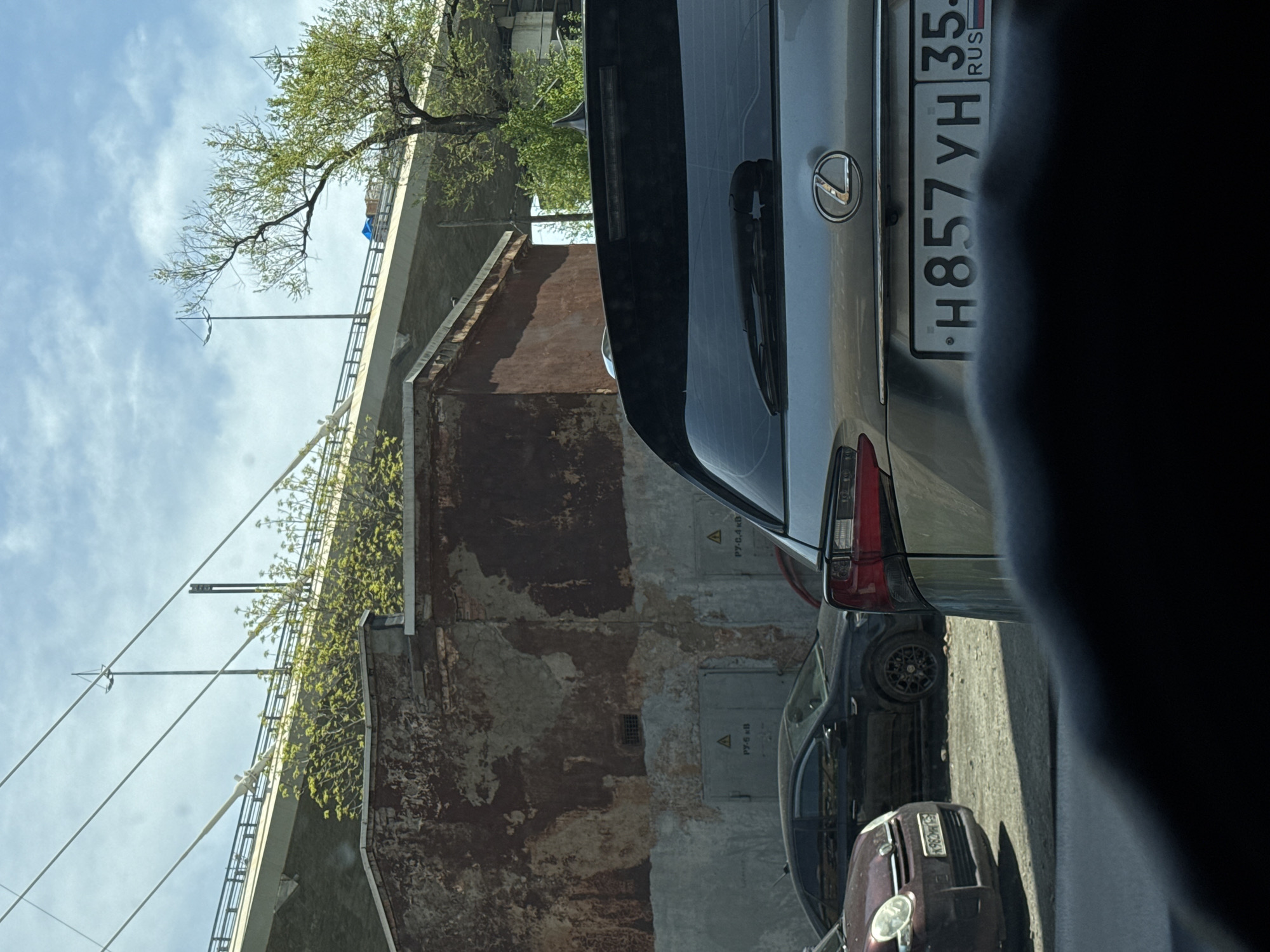 The image shows a street scene in Vladivostok, Russia. A silver Lexus SUV is partially visible in the foreground, with the license plate "H857YH 35 RUS." Parked behind the Lexus are two other cars, a dark-colored sedan, and a maroon hatchback. An old brick building with a peeling facade is located near the cars. Above the building is a bridge with green vegetation growing on it. There are power lines running parallel to the bridge. The sky is visible in the background, with some clouds.