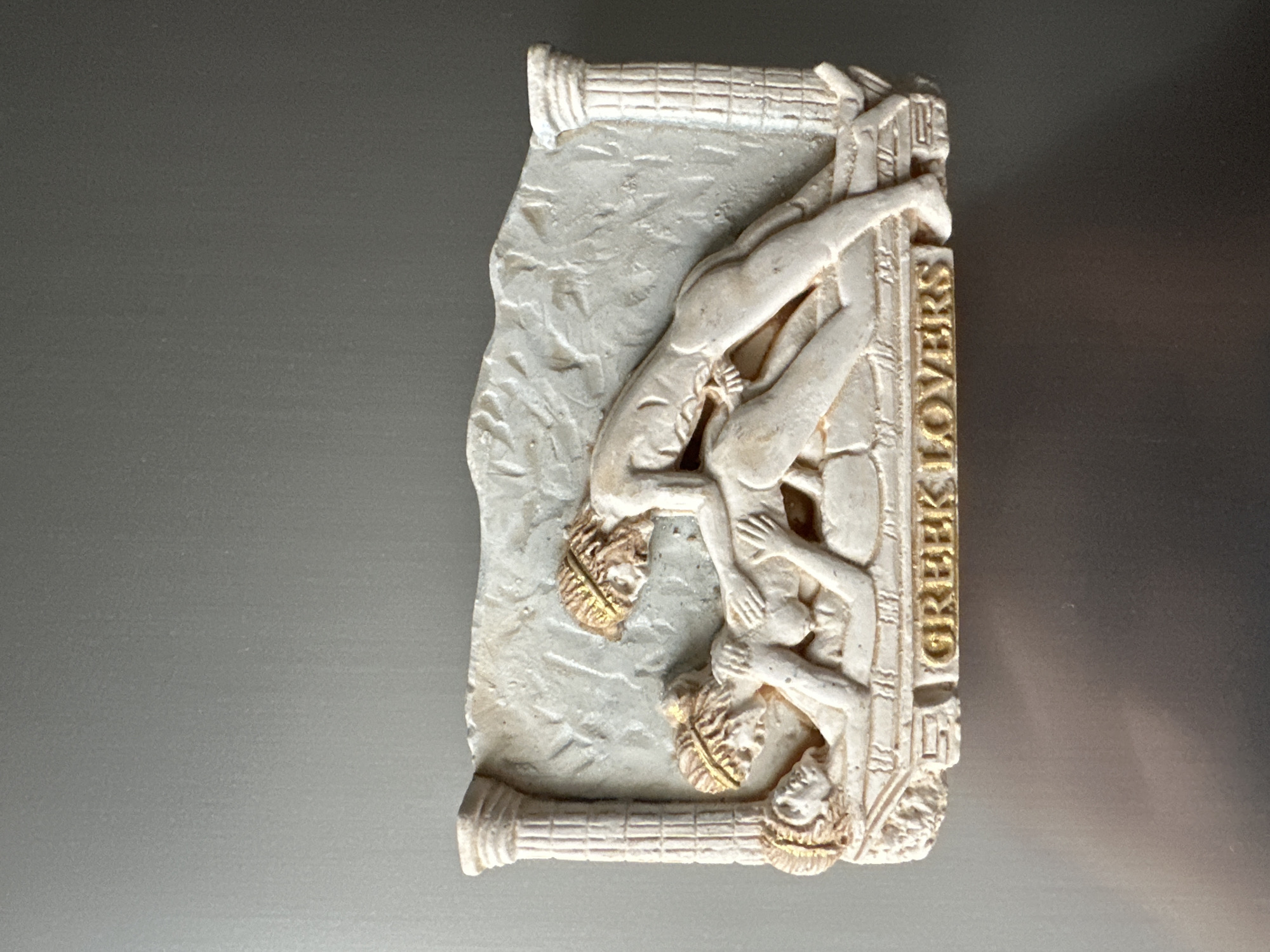 A close-up, top-down shot shows a bas-relief plaque depicting three figures in a sexual act. The plaque, with a faux marble finish, is adorned with golden accents on the figures' headbands and the text "GREEK LOVERS" at the bottom. The scene is set against a plain, grey background.