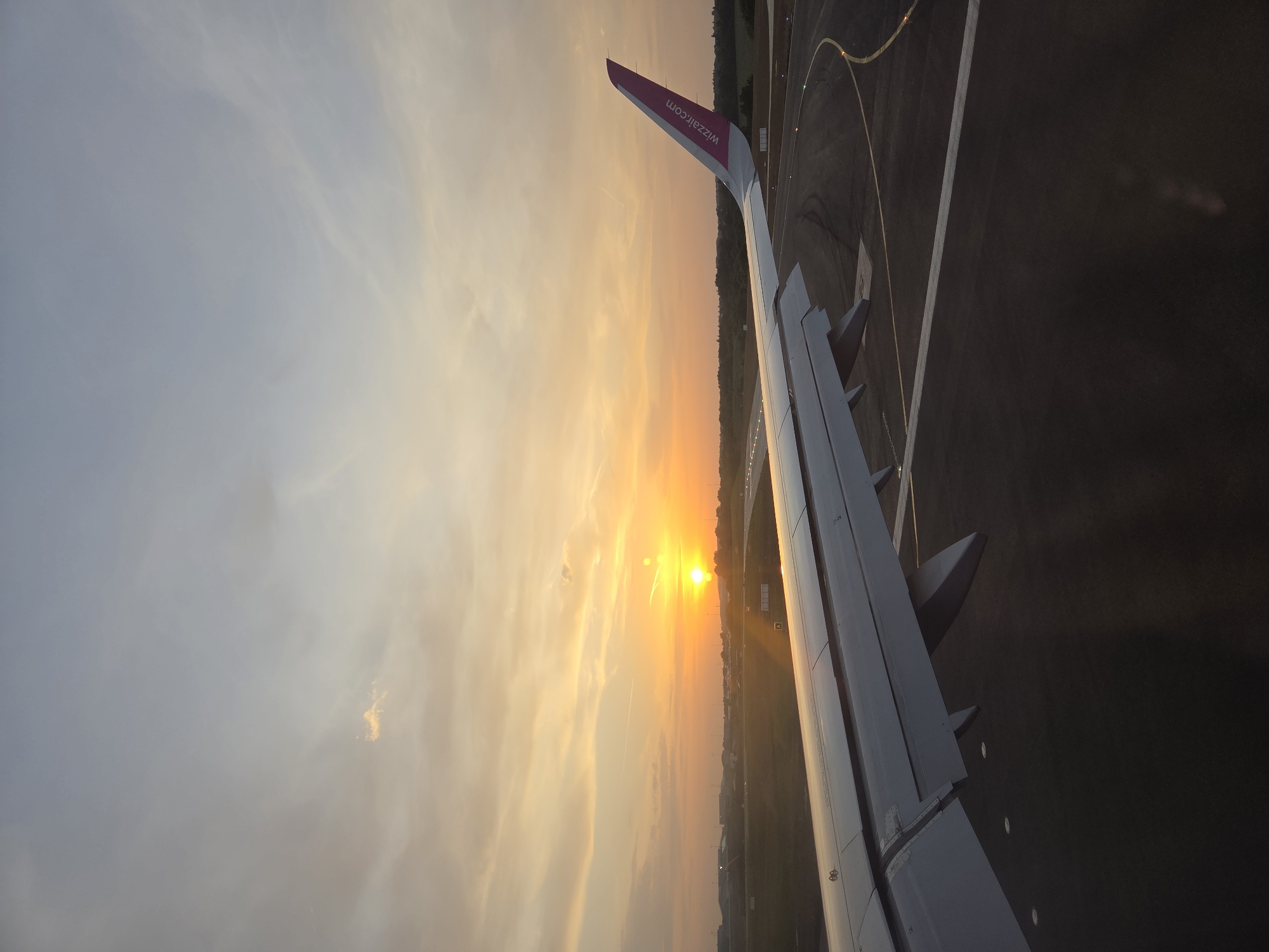 The image shows an airplane wing with the "wizzair.com" logo, presumably taken from inside the aircraft. The plane is on the runway, and the sun is setting in the background, with its light reflecting on the wing. The runway is visible below the wing. The image was not taken in North Hertfordshire, United Kingdom because it contains a runway.