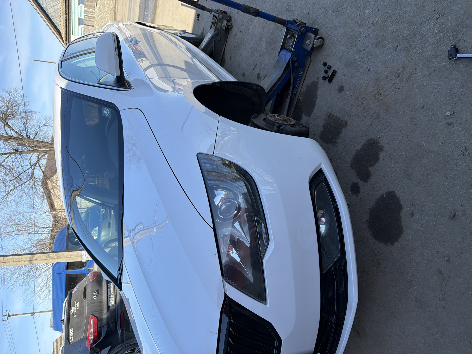 The image shows a white Skoda car being repaired outdoors. The car is raised on two blue Rossvik hydraulic jacks, with the front right wheel removed. Lug nuts and a wrench lie on the concrete pavement beside the car. There are oil stains on the ground. In the background, partially visible behind the Skoda, are a dark-colored SUV (possibly a Hyundai, based on the logo visible) and a blue van. Street elements such as a utility pole and leafless trees are also in the background, indicating an outdoor setting on a sunny day. The location is Chaikovsky, Russia.