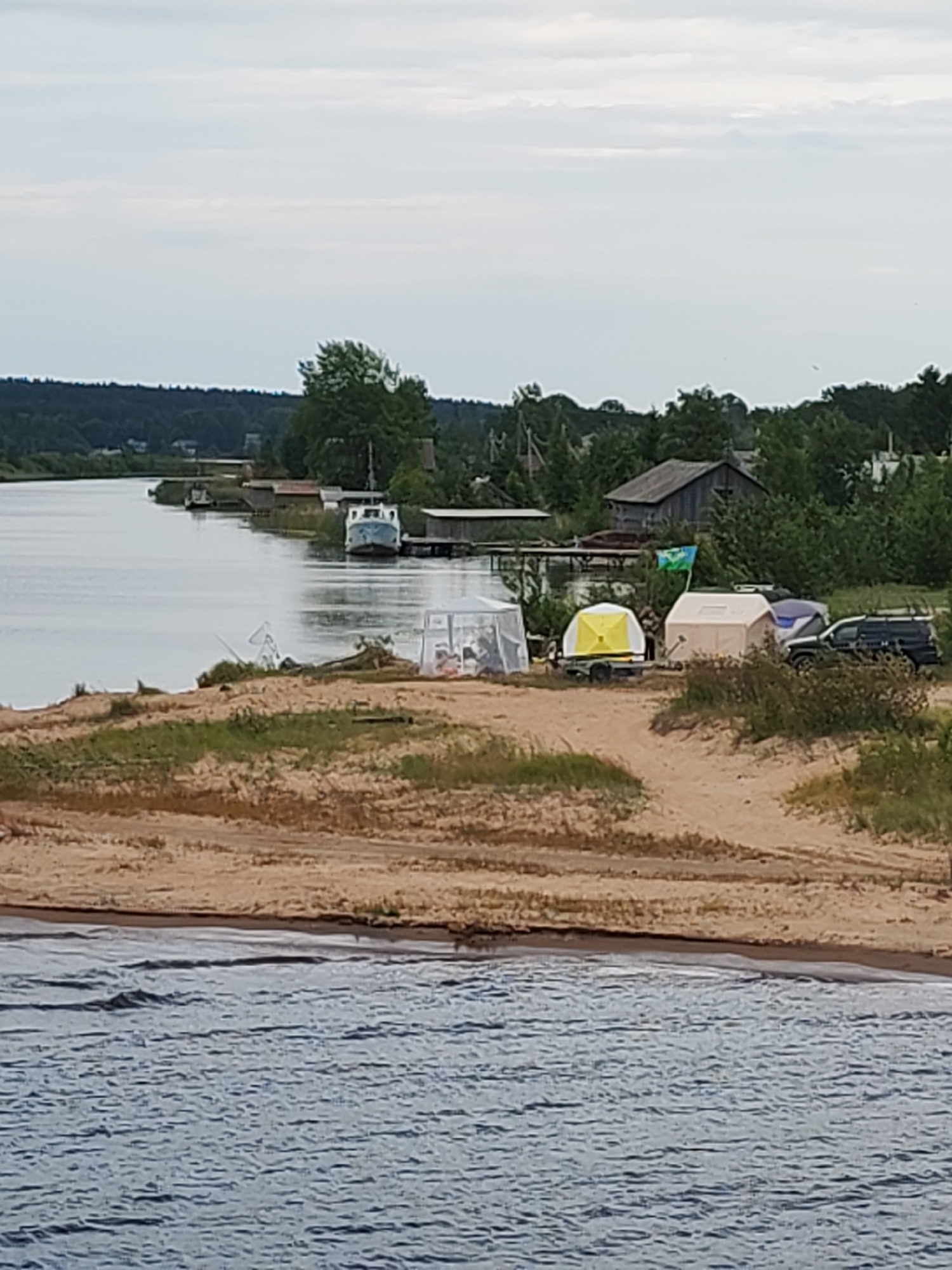 A tranquil river scene unfolds in Shalsky, Russia, under an overcast sky. The foreground is dominated by the calm, rippling surface of a wide river, its blue waters reflecting the muted light. On the right bank, a sandy embankment gives way to a grassy area where a campsite is set up. Several tents are visible, including a prominent white tent and a smaller yellow-topped tent on a trailer. A dark-colored SUV is parked nearby, and a blue and green flag flutters from a pole. Behind the campsite, a cluster of trees and simple wooden structures suggest a small settlement or rural area. Across the river, the opposite bank is lined with trees, and a few more wooden buildings and a boat are visible near the water's edge. The overall impression is one of a peaceful, natural setting with a hint of human activity.