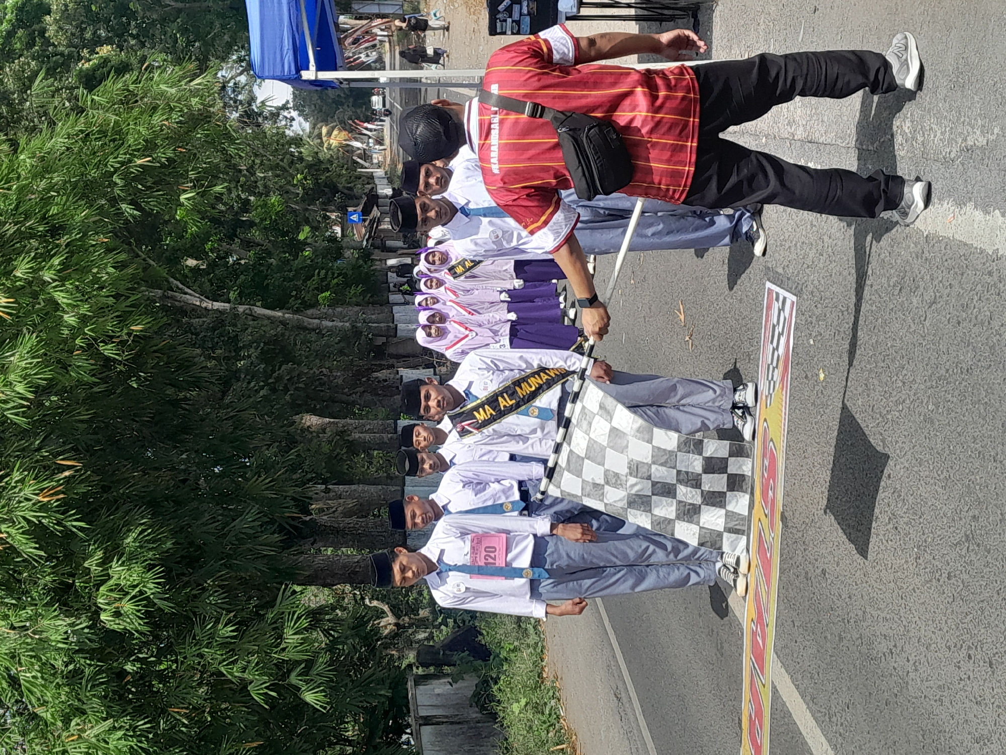 In Karangsari, Indonesia, on a paved road lined with trees and residential structures, a group of students in white shirts, blue ties, and black peci hats stand at attention. One student, identified by bib number 120, holds a checkered flag at the finish line, suggesting a race event. Behind him, other students and a person in a red and yellow striped shirt holding a fanny pack are also present. The scene is illuminated by sunlight, creating distinct shadows on the asphalt. A blue canopy tent is visible in the background, along with other race participants and spectators.