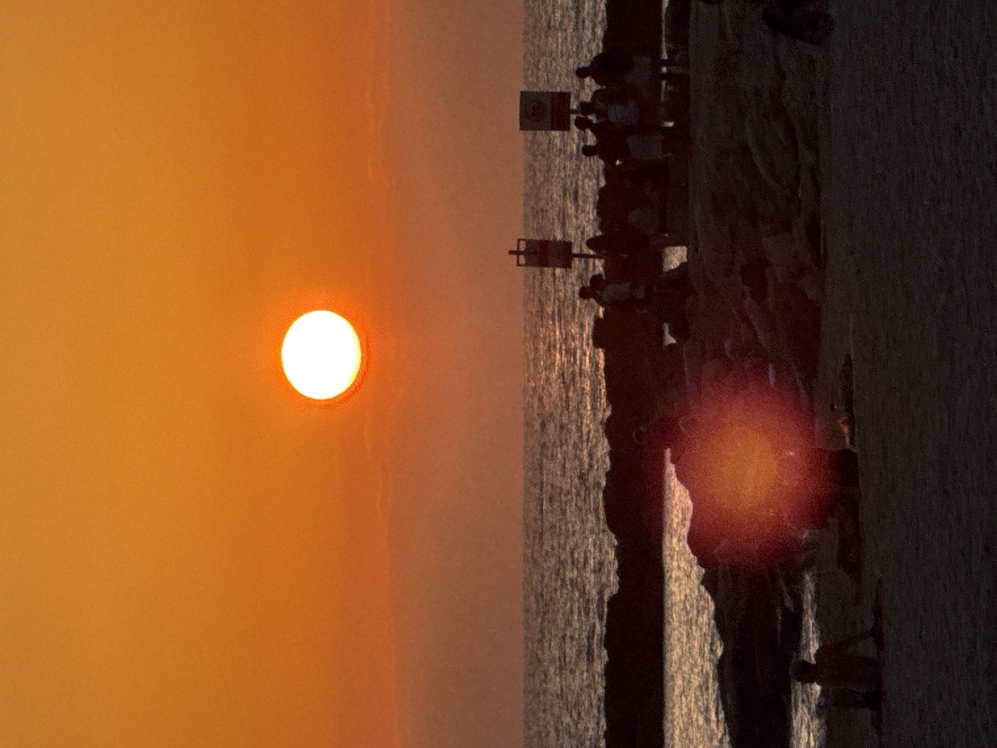 The image depicts a sunset over the Mediterranean Sea, likely in Tel Aviv, Israel, given the characteristic urban coastline. The sun, a brilliant white orb with a soft orange halo, is positioned in the upper center of the frame, casting a warm, vibrant orange glow across the sky and the water. The sea exhibits choppy waves, reflecting the sunset's light.
In the foreground, a sandy beach is visible, transitioning into darker, rocky areas along the shoreline. Several silhouetted figures are scattered across the scene. On the beach in the lower left, a lone individual sits facing away from the camera. Further up the beach, near the rocky coastline, a group of people are gathered, their forms indistinct in the low light. Two signs on poles are visible amidst the group, their content obscured by distance and shadow. The overall atmosphere is serene, capturing a moment of twilight by the sea.