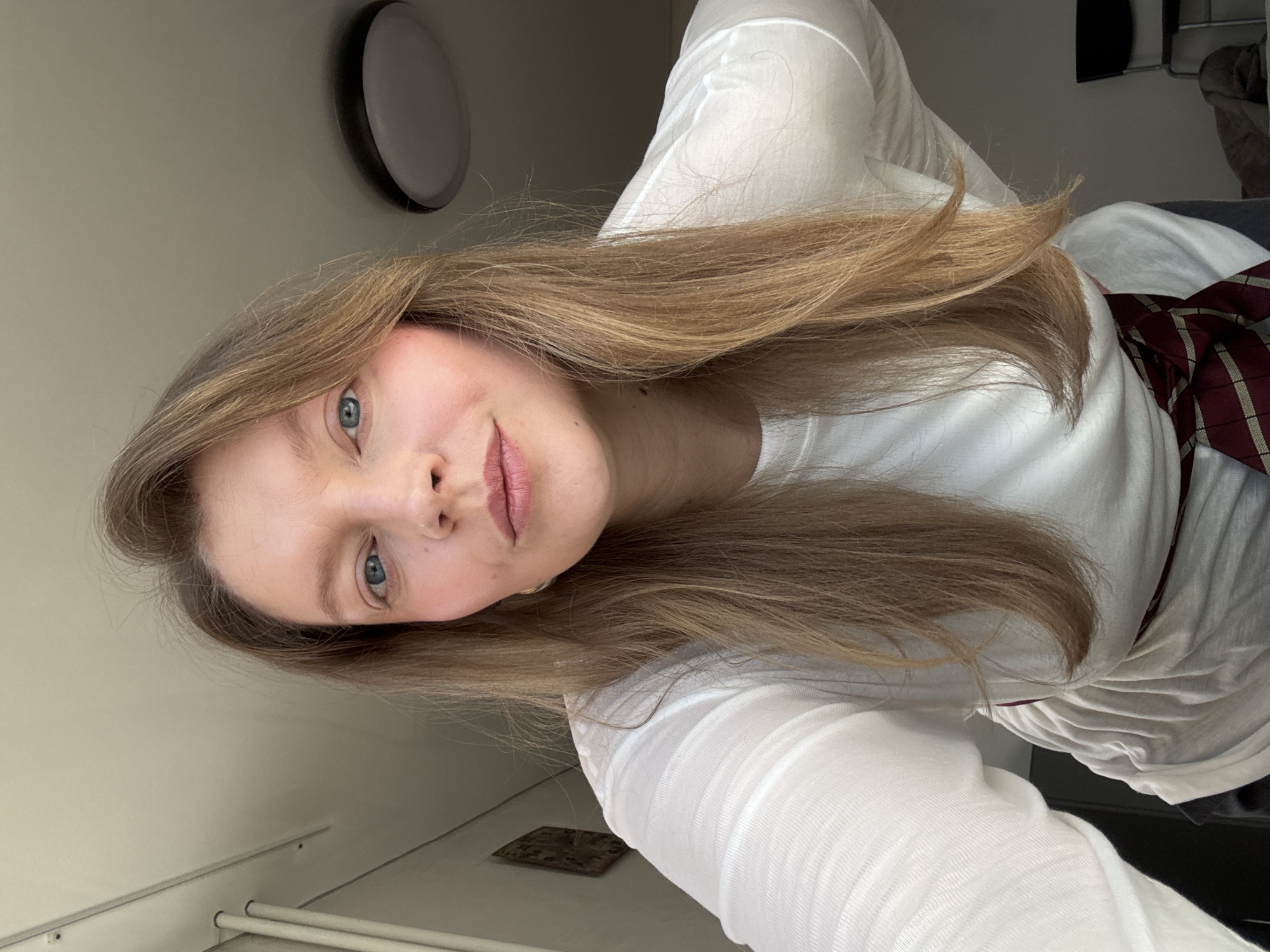 A woman with long blonde hair and blue eyes is posing for a selfie. She is wearing a white long-sleeved shirt and a tie. The background includes a light fixture on the wall and part of a window. There is no information to indicate this photo was taken in Izhevsk, Russia.