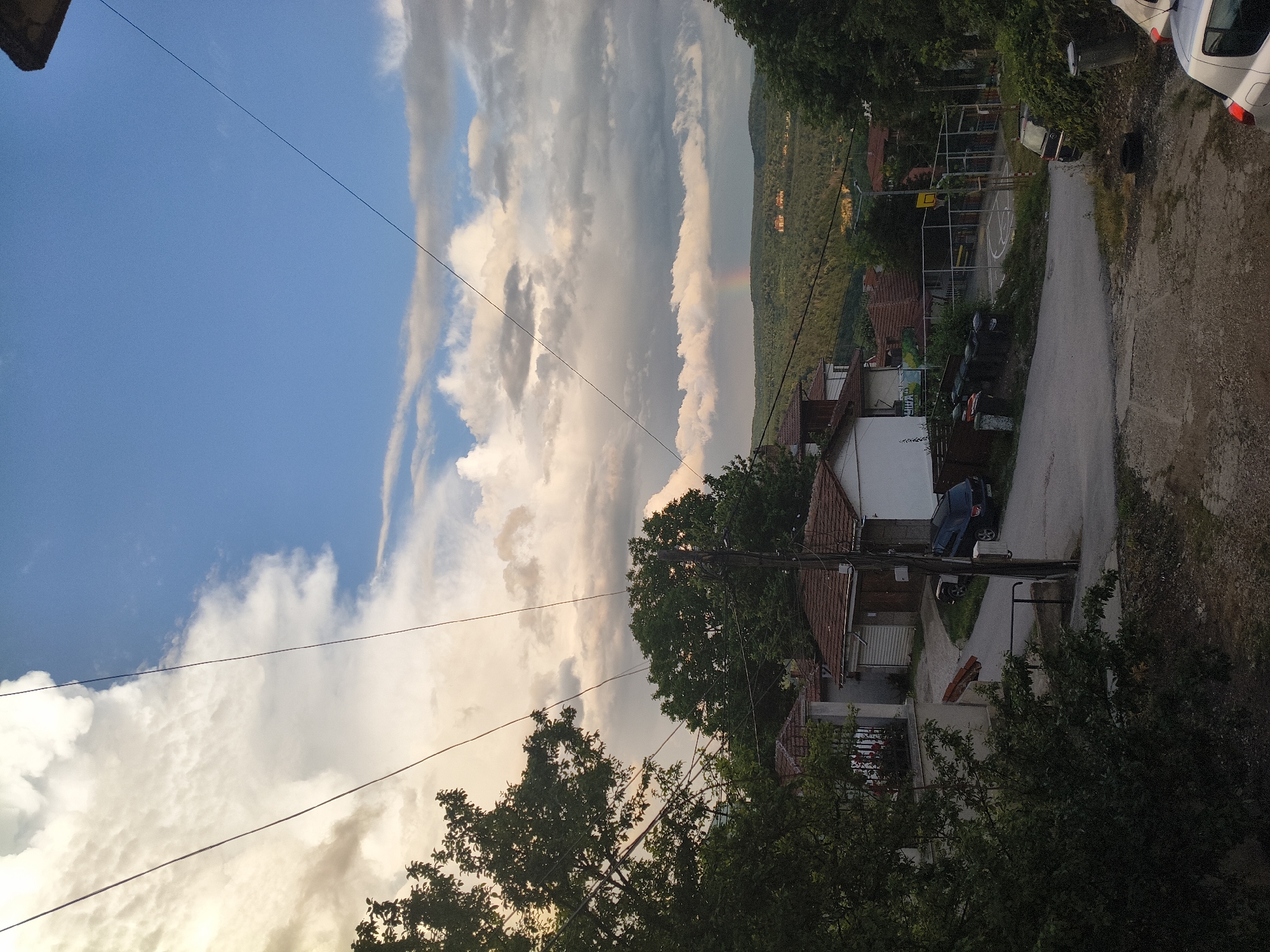 The image captures a panoramic view of a residential area, likely in the late afternoon or early evening, bathed in soft, warm light. The sky dominates the upper portion, showcasing a dramatic expanse of fluffy cumulus clouds, ranging from bright white to soft grey and pinkish hues, contrasting with patches of clear blue. A faint rainbow is visible high over a distant, verdant hillside, hinting at recent rain.
In the mid-ground, nestled among abundant green trees and foliage, are several houses with traditional tiled roofs, some with white facades. A narrow, unpaved road or path winds through the scene, connecting to a more defined dirt track in the foreground. A white car is partially visible parked at the upper right, and a blue car is parked near one of the houses in the mid-ground. A small, enclosed basketball court with a yellow hoop and marked lines is a prominent feature, suggesting a community space.
Numerous power lines or utility wires crisscross the sky, adding an urban element to the otherwise natural landscape. The foreground is framed by dark green trees and bushes. On a partially obscured white sign near one of the houses, some Cyrillic text is visible, beginning with "СПИРА..." The overall setting depicts a quiet, residential neighborhood on the outskirts of a town, consistent with the provided location context of Veliko Tarnovo, Bulgaria.