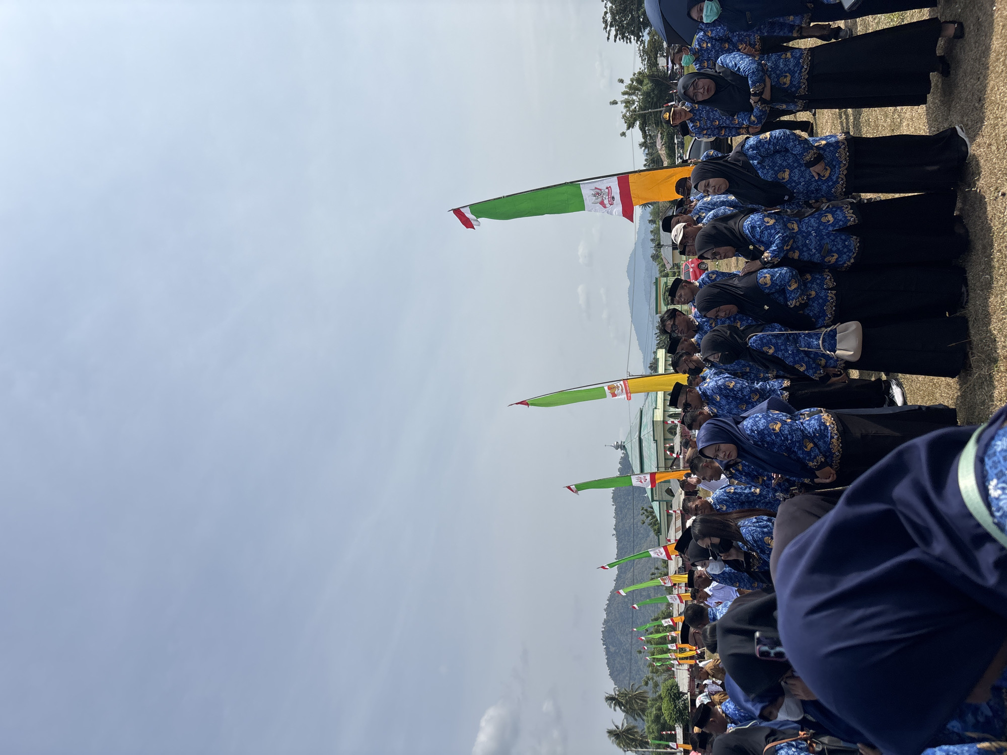 The image shows a group of people standing in an outdoor field, possibly for an event. Many individuals are wearing matching blue uniforms with a patterned design, and several women are wearing headscarves. In the background, there are several tall flags with green, yellow, and red colors, some featuring emblems. A mosque-like building with a green dome is visible behind the flags, and a hill with trees can be seen in the distance. The sky is partially cloudy. The scene is likely in Torosiaje, Indonesia, given the provided context.