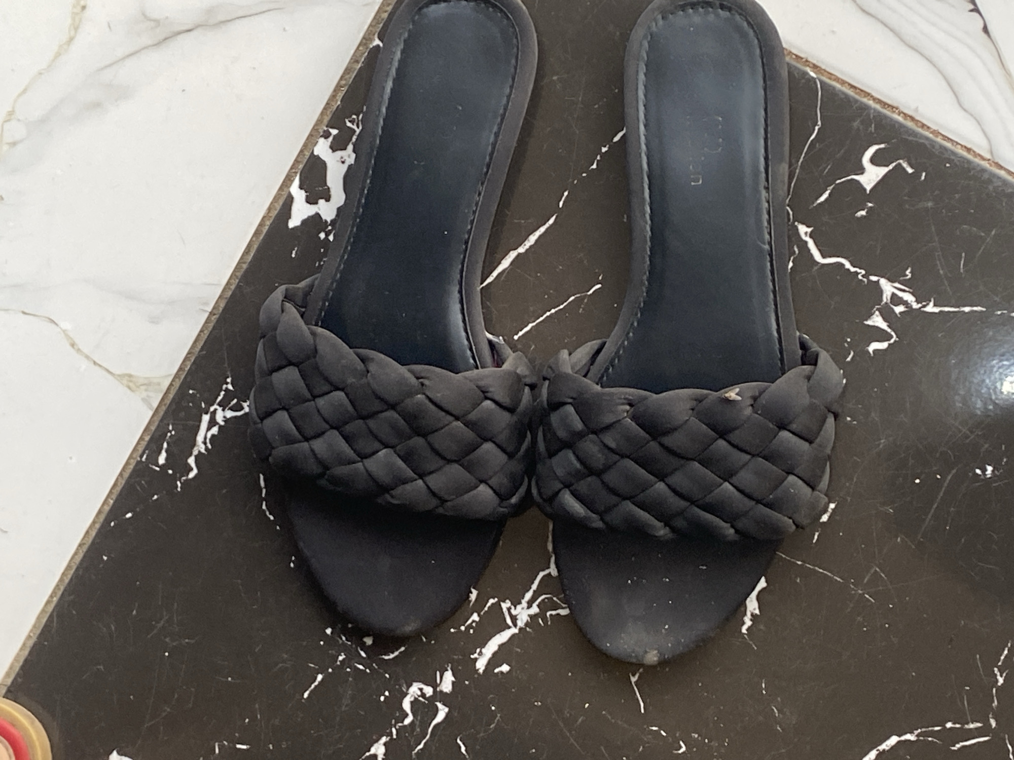 Two black slide sandals with braided straps are positioned diagonally on a dark, marble-patterned surface, which is placed on top of a lighter, marble-patterned surface. The sandals are side-by-side, with the soles facing upwards. The braided straps are made of a soft material and are woven in a diamond pattern. The footbeds of the sandals appear to be made of a darker material, and there is faint branding visible on the right sandal's footbed. The scene is lit from above, casting subtle shadows. There are no people present in the image. The location is Jauro Yinu, Nigeria.