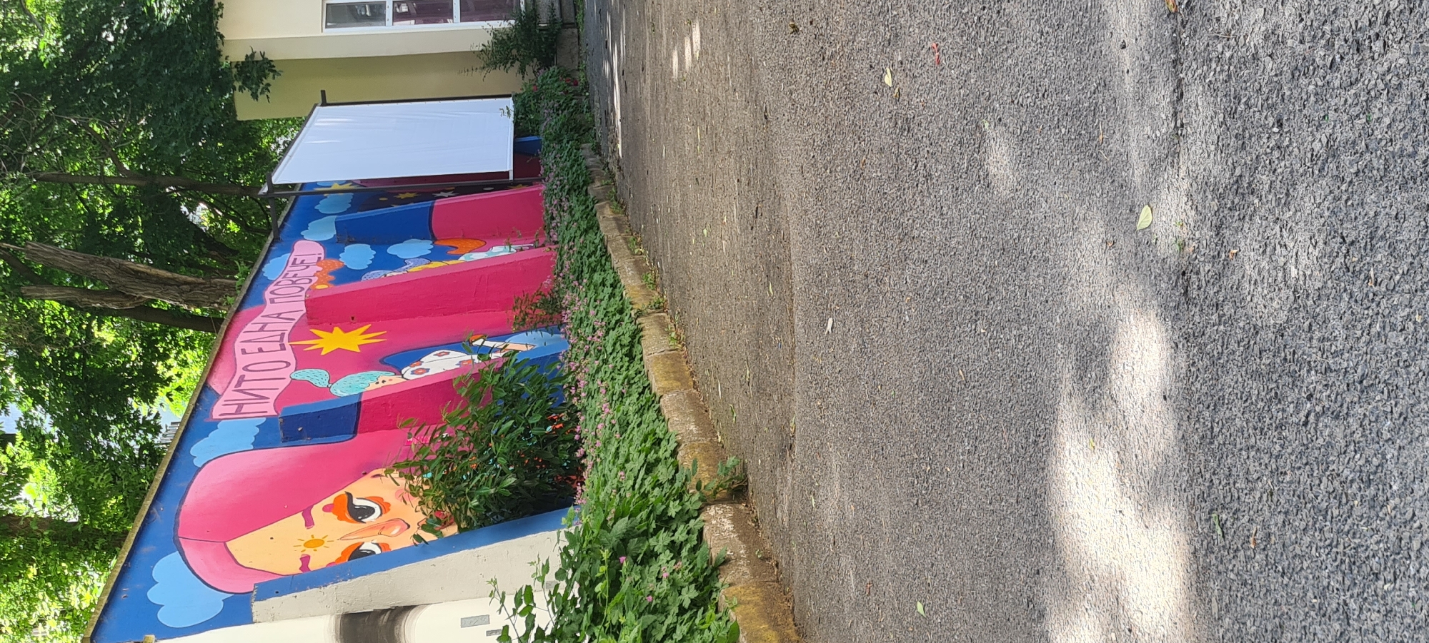 The image shows a section of street in Sofia, Bulgaria, next to a low wall with a colorful mural painted on it. The mural depicts a face with pink hair and other whimsical images. The words "НИТО ЕДНА ПОВЕЧЕ!" are written on a banner in the mural. A small bush grows in front of the mural wall. The street is asphalt and has patches of sunlight and shade. A white screen is visible behind the wall. Trees are also visible in the background.