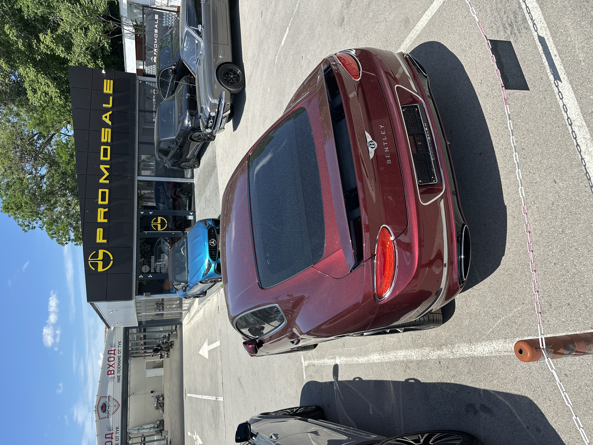 The image shows a burgundy Bentley parked in a lot with "PROMOSALE" and "PORT MOTORS" signage visible, suggesting it is a car dealership. The background includes other cars of different makes and colors. The setting is likely an outdoor parking area associated with a commercial business. The OCR indicates that "PROMOSALE" is a dealership with signs in both English and Bulgarian, including phrases like "ПРОДАЖБА" (Sales) and "BHOC" (Import). The address is likely located in Varna, Bulgaria, as suggested by the location description of "НИЕ ПОЕМАМЕ ОТ ТУК" (we pick up from here). The parking area is marked with white lines and a chain. The weather appears sunny, judging by the shadows.