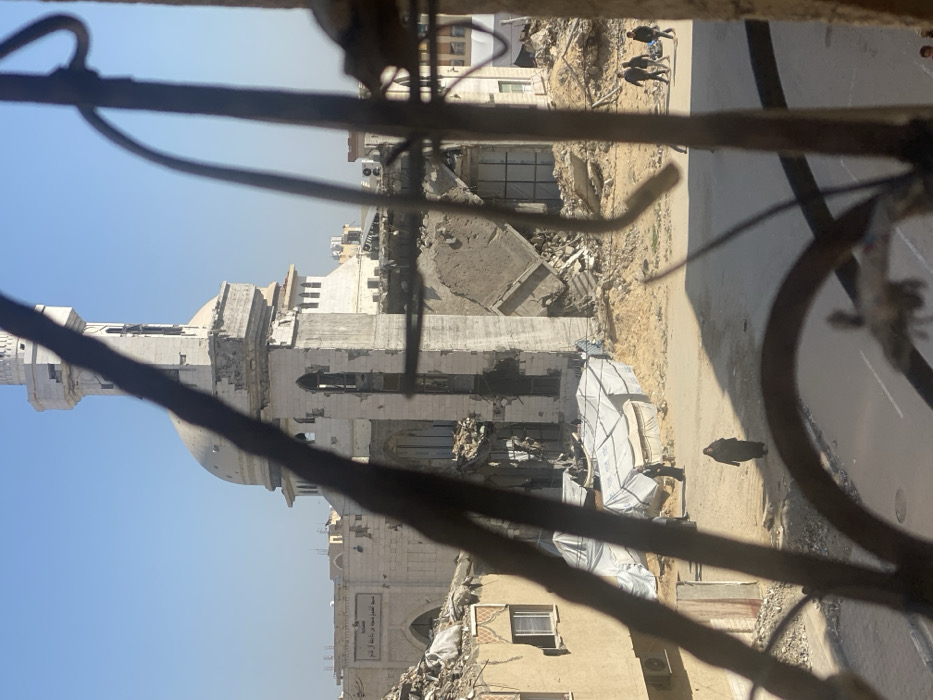 The image depicts a scene of extensive urban destruction in Khan Yunis, Palestinian Territory, viewed through a set of heavily corroded and bent metal bars in the foreground. The most prominent structure is a heavily damaged mosque, identified by an Arabic sign on an adjacent building as "مسجد المحمود بن جبير بن ثابت" (Al-Mahmoud bin Jubeir bin Thabit Mosque). Its white minaret is partially demolished, showing holes and missing sections, while the main dome structure is also visibly damaged. The mosque's lower sections exhibit significant structural collapse, with large portions of walls missing and interior debris visible.
Surrounding the mosque and extending across the midground is a vast expanse of rubble and demolition debris, consisting of broken concrete, rebar, and pulverized building materials. Several partially destroyed buildings are visible in the background and to the left, some reduced to skeletal frames. In the midground, to the left of the mosque, several white tarp-covered makeshift structures are erected amidst the debris on sandy ground.
A paved road runs horizontally across the lower part of the scene. On this road, several individuals are walking: one person in a dark abaya walks alone from left to right in the foreground, and a group of three individuals, also in dark clothing, walks further down the road to the right, with one appearing to carry an item. Another person in dark attire is visible near the temporary tarp structures. The sky above is clear blue, indicating daytime conditions. Shadows are long, suggesting morning or late afternoon.