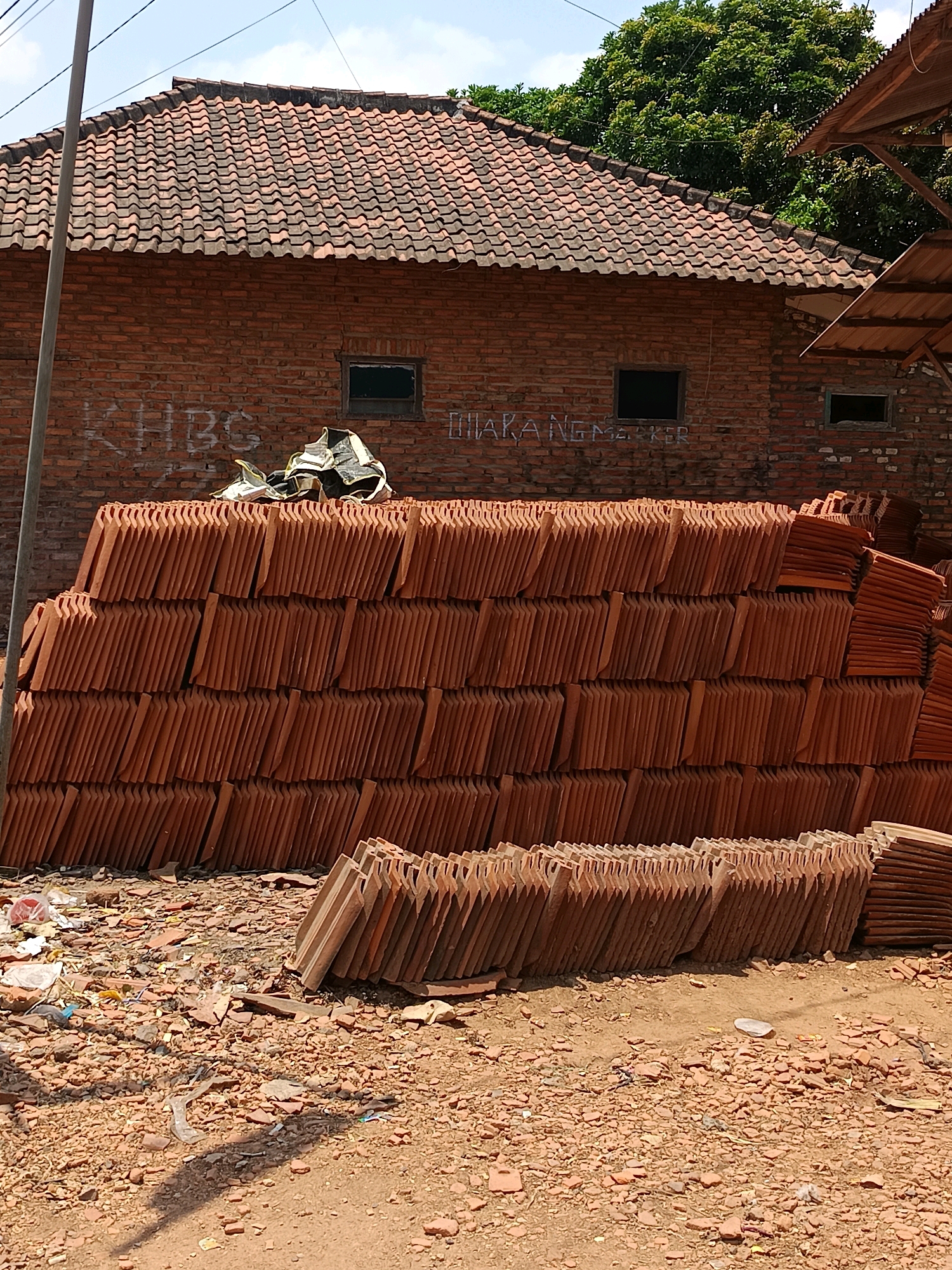 The image captures an outdoor scene in Blega, Indonesia, dominated by a stack of reddish-brown roof tiles. The tiles are neatly piled, creating a textured wall that occupies a significant portion of the frame. Behind the tiles, a brick building with a tiled roof is visible. Graffiti, appearing as white spray paint, is present on the brick wall, with two windows visible above the tile stack. A large, mature tree with dense green foliage is situated to the right of the building. The ground in front of the tiles is covered with dirt, small rocks, and scattered debris, suggesting a construction or storage area. The sky above is a clear blue with scattered white clouds. No people are present in the image.