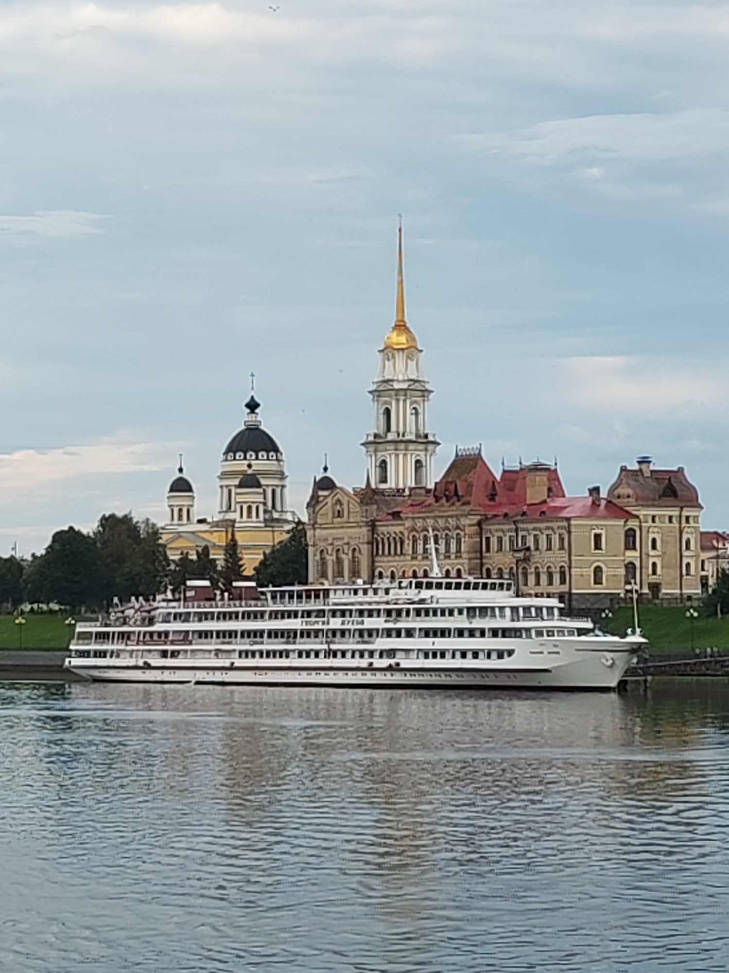 A long, white cruise ship is docked on a wide, calm body of water. The ship has multiple decks, many windows, and a prominent white hull with red trim. Behind the ship, on the shore, is a complex of buildings with distinctive architecture. One building is a large yellow structure with a black dome and cross, resembling a church or cathedral. Adjacent to it is a taller, white bell tower with a gold spire. Another building complex to the right features red tiled roofs and beige walls with numerous windows, suggesting a historical or governmental purpose. Lush green trees and grass are visible on the embankment behind the buildings. The sky is overcast with scattered clouds. The overall scene depicts a serene riverfront vista with a cruise ship and historic architecture in the city of Bol'shie Mkhi, Russia.