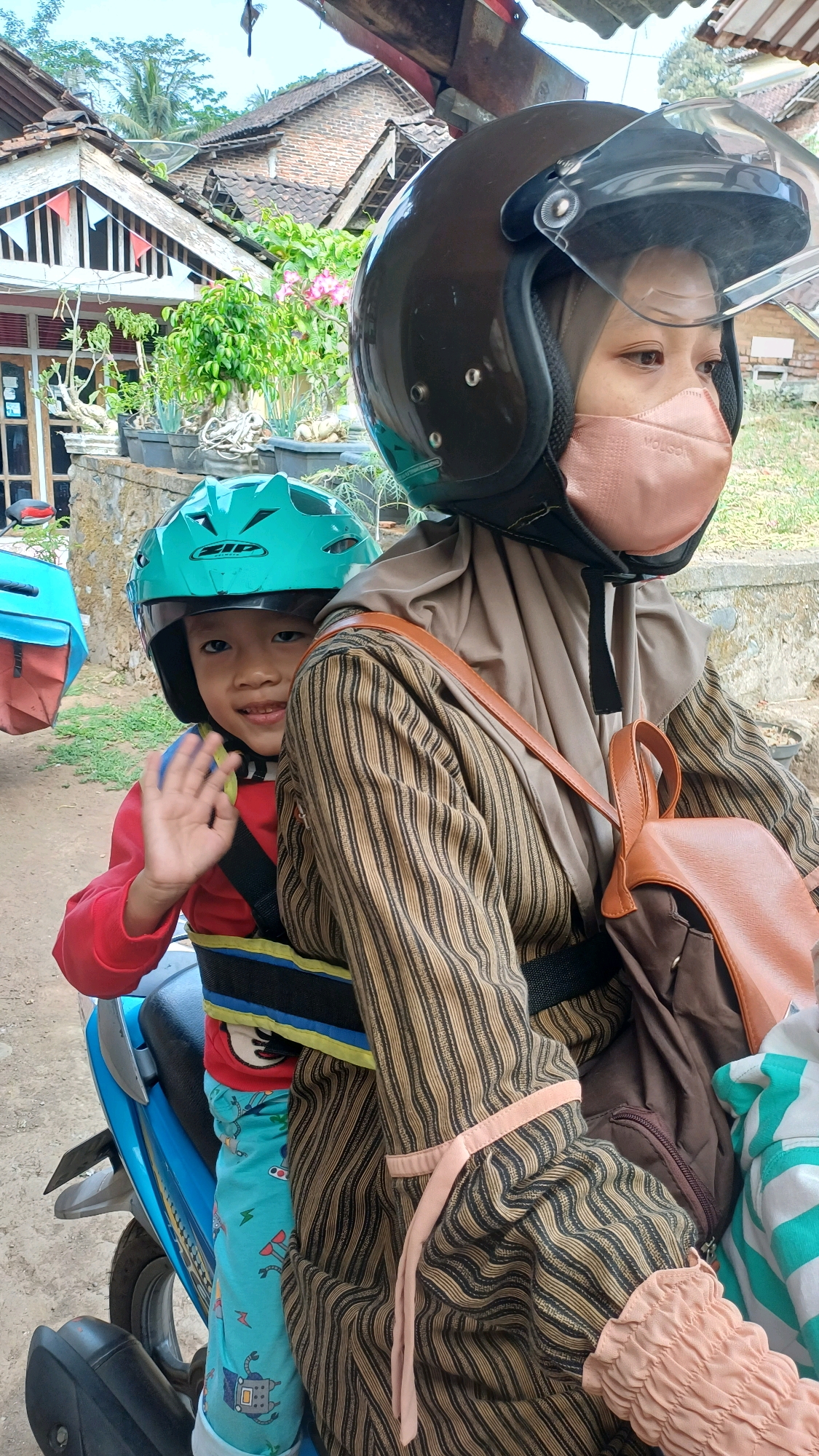 In Wonokerso, Indonesia, a young boy wearing a teal helmet and a red shirt is seated on the back of a blue motorcycle, facing forward. He is waving to the camera with his right hand. A woman, also wearing a helmet and a pink face mask, is seated in front of him, driving the motorcycle. She has on a brown and black striped outfit and a light brown backpack. The background shows a rural Indonesian setting with traditional houses and lush greenery.