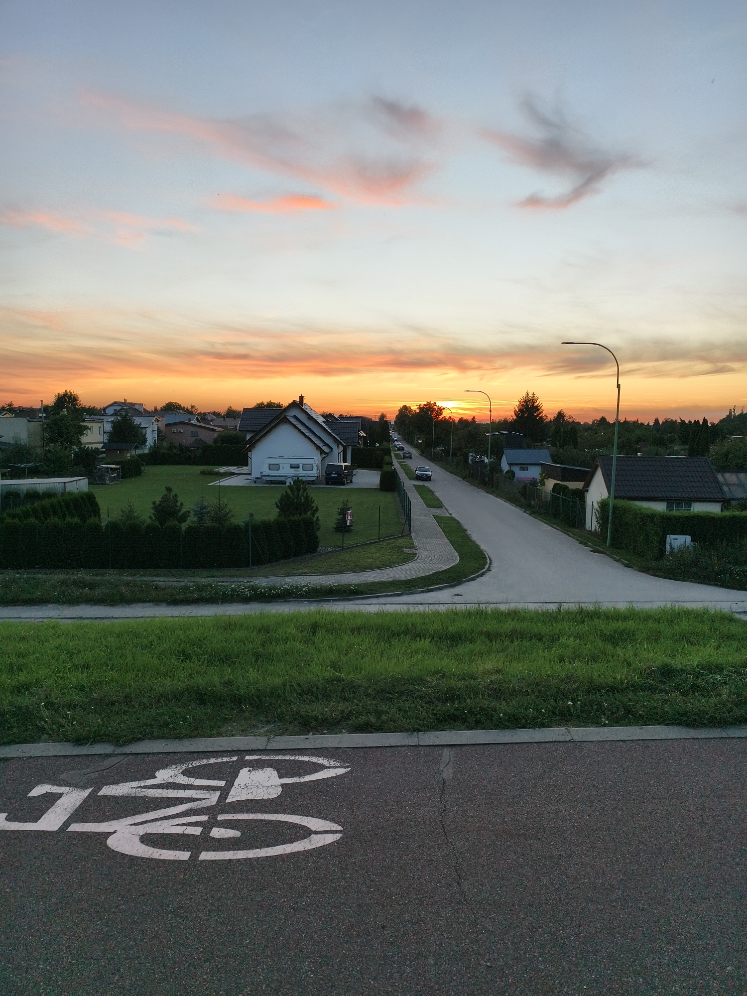 The image shows a street scene at sunset in Tczew, Poland. The sky is filled with orange and pink clouds. In the foreground, there is a bicycle lane marking on the asphalt. The street curves to the right, with houses and trees lining both sides. There are a few cars parked along the street and some garden fences. The overall impression is a quiet suburban evening.