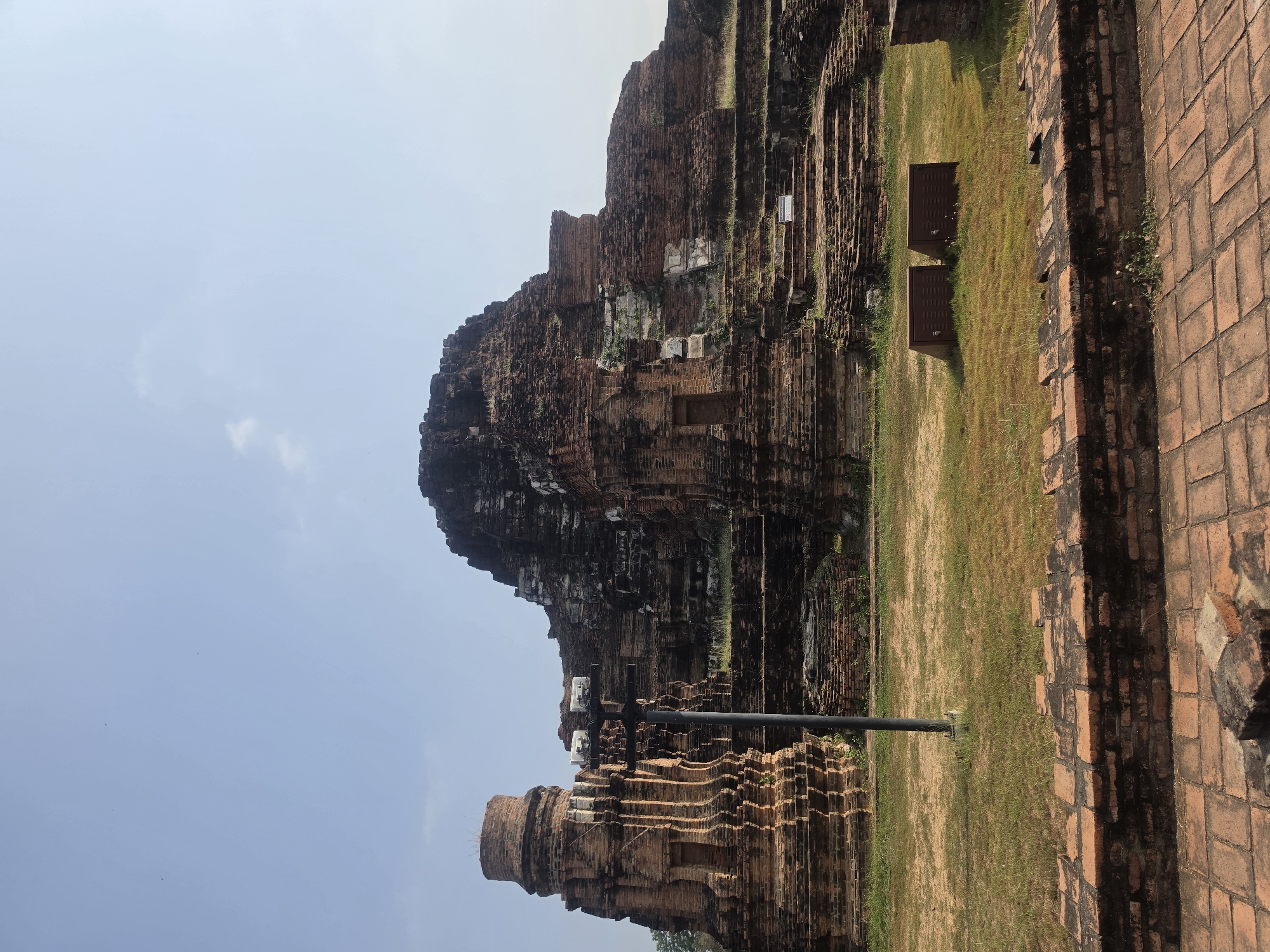 The image depicts ancient brick ruins in Phra Nakhon Si Ayutthaya City Municipality, Thailand. The ruins consist of a large, layered brick structure partially collapsed, set against a blue sky. There is a smaller brick structure in the foreground and grassy area.