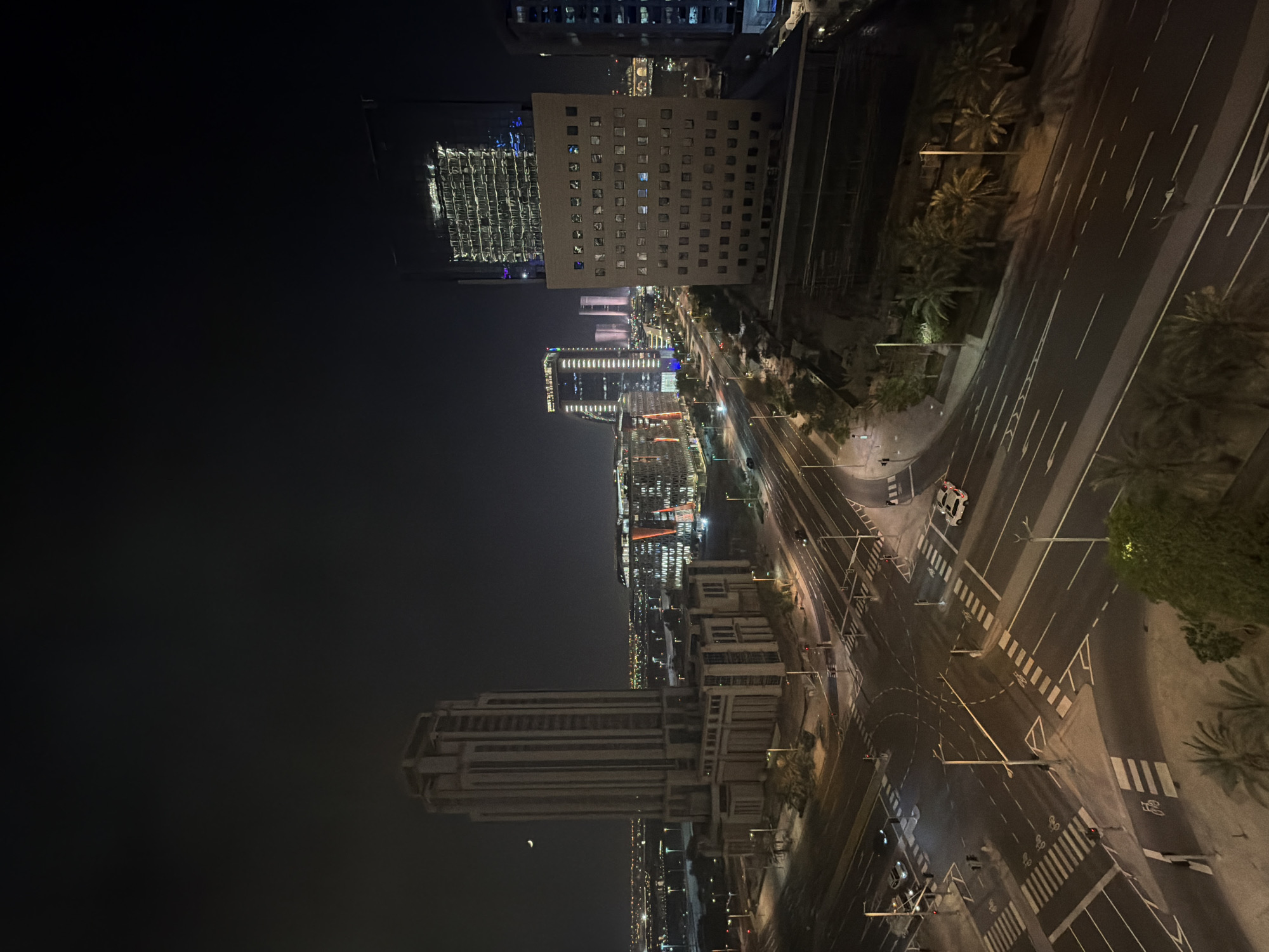 This is an aerial night photograph of a modern cityscape with multiple tall buildings and a complex intersection of roads. The sky is dark, with a faint crescent moon visible in the upper left quadrant of the image. The buildings are illuminated by interior and exterior lights, creating a pattern of illuminated windows and architectural features.
The most prominent building in the foreground, on the left, is a tall, light-colored, rectangular skyscraper with a stepped top. It has many windows and a solid, layered base structure. To the right of this building, and slightly further back, is another skyscraper with a glass facade that reflects the city lights. In the middle ground, several other modern buildings are visible, some with illuminated vertical accents or patterned facades. The furthest buildings in the background are less distinct and appear smaller and more numerous, suggesting a sprawling urban area.
A multi-lane road intersection with pedestrian crossings dominates the lower half of the image. Traffic lights are visible, and a white police car is stopped at the intersection. Several other vehicles are on the roads, appearing as streaks of light or distinct shapes. The roads are marked with white lines, including crosswalks and directional arrows. Palm trees are interspersed along the sides of the roads and in some of the landscaped areas between buildings. The overall scene conveys a sense of a vibrant, active urban environment at night.