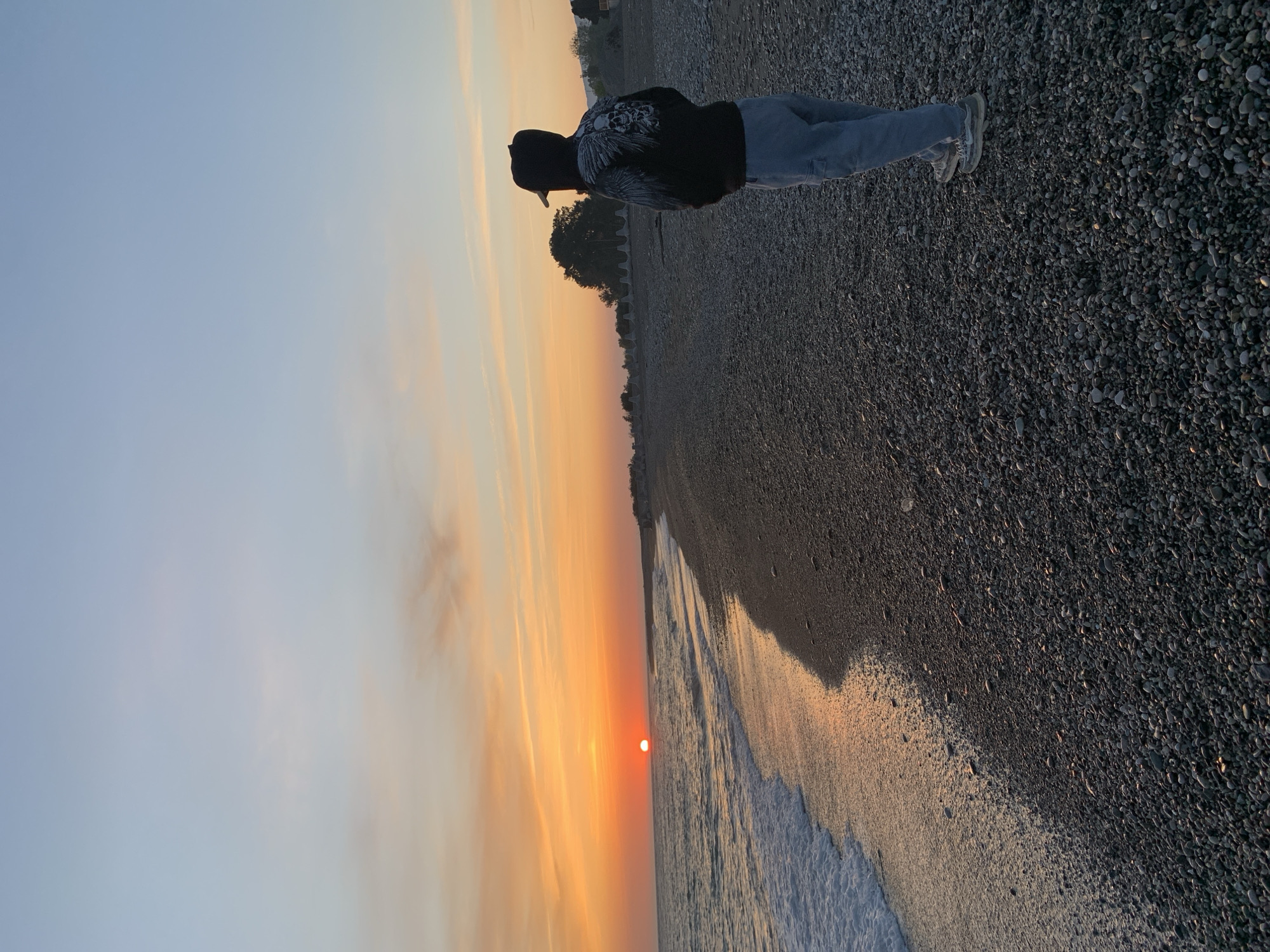 A medium, rotated, outdoor shot depicts a person standing on a pebble beach with the sun setting over the water. The person is wearing a black hoodie with a graphic on the back, light blue jeans, white shoes, and a black baseball cap. They are facing the ocean and appear to be looking at the sunset. The beach is composed of small, dark-colored pebbles. The sea is calm with small waves breaking on the shore. The sky is a gradient of light blue to orange, with the setting sun visible as a bright orange orb near the horizon. The photo was taken at the beach, in the city of Inkiti, Abkhazia.