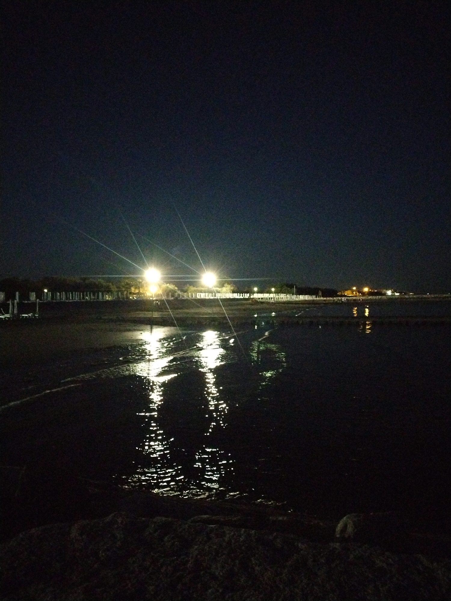 The image depicts a nighttime scene at a beach in Caorle, Italy. Two bright artificial lights, likely streetlamps or stadium lights, are prominently visible in the mid-ground, casting strong, starburst-like beams across the dark sky. The lights are positioned near a line of trees or vegetation. In the foreground, a body of water, possibly the sea or a shallow lagoon, reflects the artificial lights, creating shimmering patterns on its surface. The ground in the immediate foreground appears to be sandy or pebbly. Structures that resemble beach chairs or loungers are faintly visible to the left. The overall atmosphere is dark and somewhat serene, with the artificial illumination providing the primary visual contrast against the night sky and dark water.