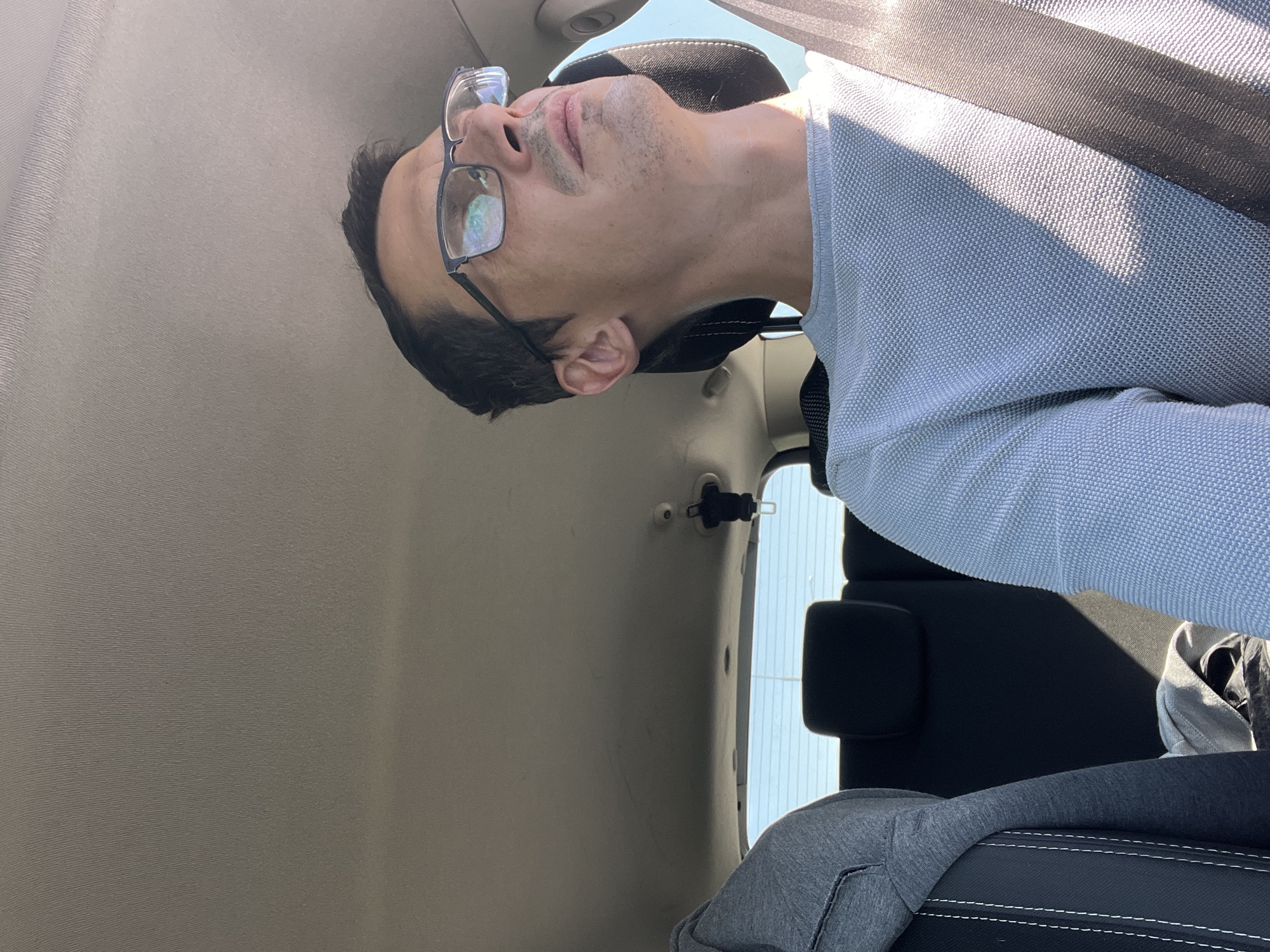 A close-up shot shows a man wearing glasses and a light blue shirt, seemingly asleep in a car. The camera angle is unusual, tilted to the side, which makes the man's head appear to be resting back. Sunlight streams in from the side, casting shadows across his face and shirt. The background reveals the interior of a car, with a beige headliner and black seats visible. A seatbelt is also present, suggesting the man is in a passenger seat. The overall impression is one of a quiet moment of rest during a car journey. The date is not visible in the image, however, we know that the location is Zabrze, Poland. No text is visible in the media.