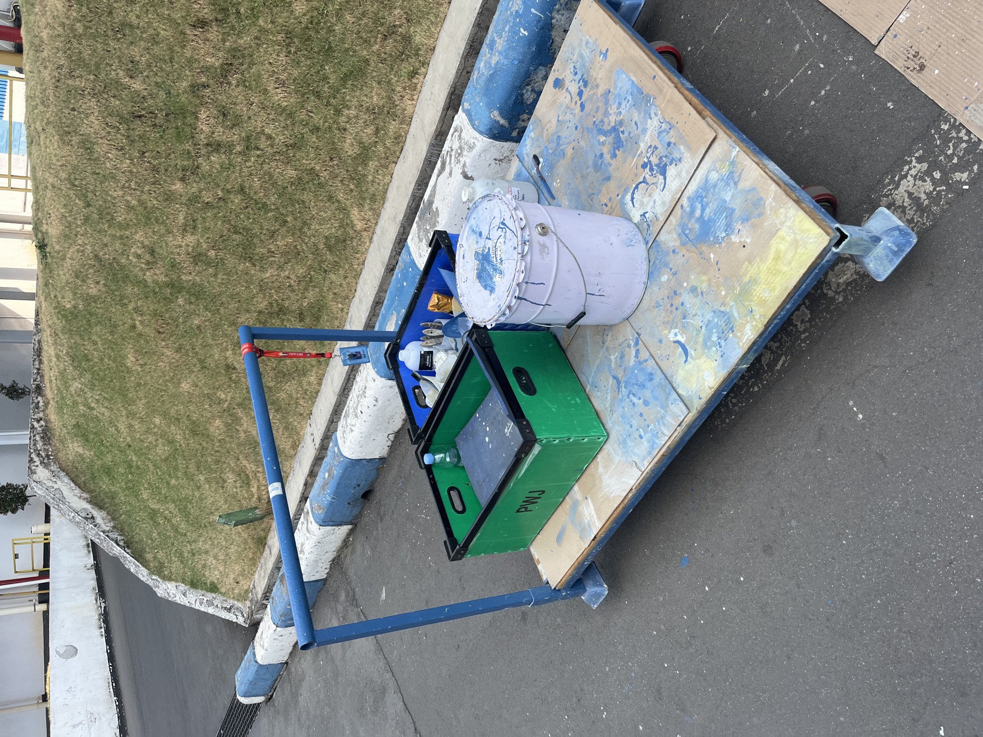 An outdoor scene in Pelang, Indonesia shows a blue cart with a wooden platform on wheels. The platform is splattered with blue and white paint. On the platform, there are two plastic bins. The green bin on the left has the letters "PWJ" printed on its side. Inside the green bin is a small bottle of water and a dark-colored object. The blue bin on the right contains several bottles and tools, including what appear to be paint brushes. Next to the blue bin is a large, light purple bucket with a metal handle, also covered in blue and white paint splatters. A white bottle with a blue label is visible behind the purple bucket. The cart is positioned on asphalt next to a grassy embankment bordered by a concrete curb painted with blue and white stripes. In the background, there are buildings and vegetation.