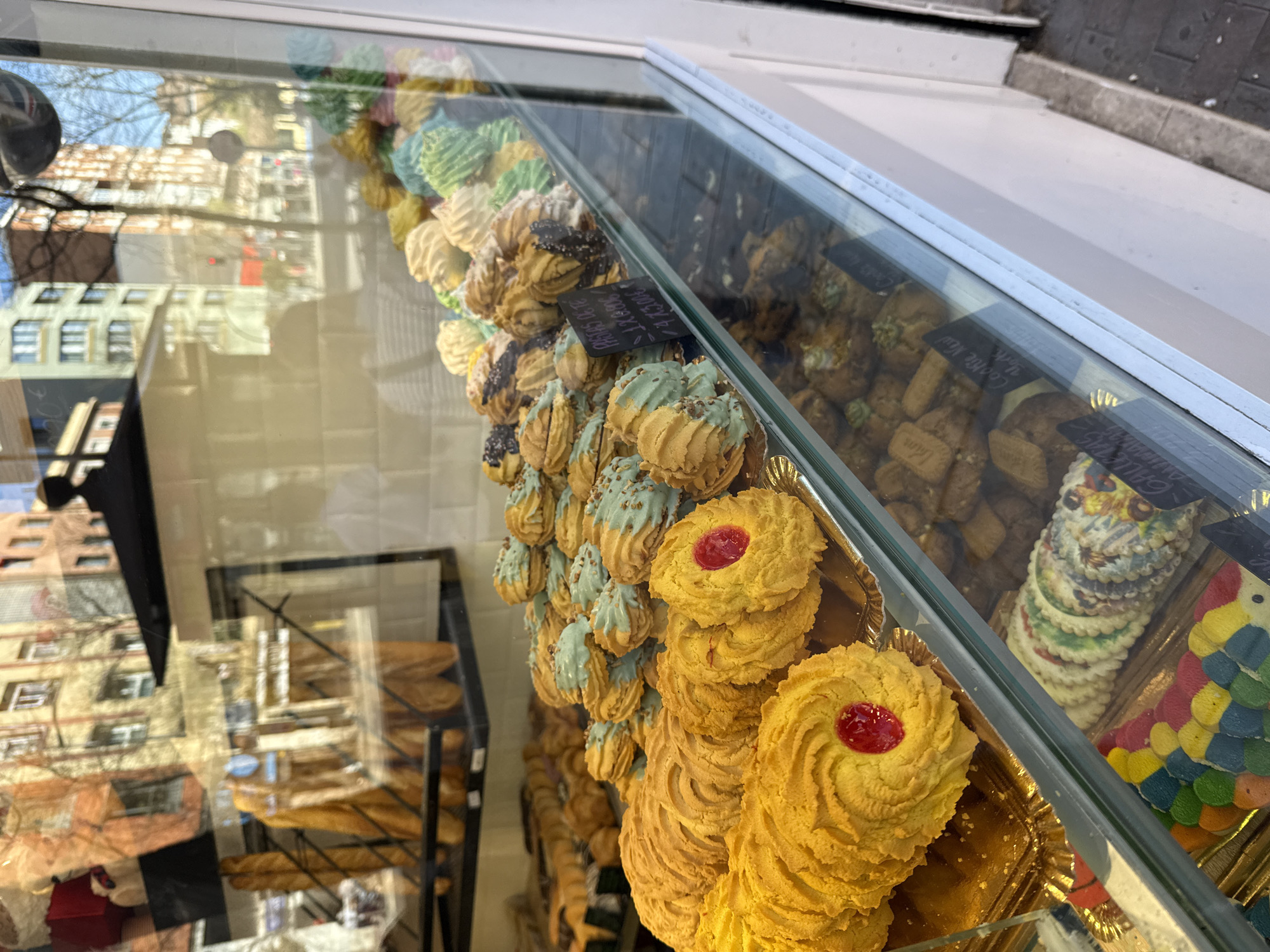 A display case in Madrid, Spain, showcases an assortment of cookies and pastries. The case contains rows of meringue-style cookies in various colors (green, white, blue) with chocolate drizzle, cookies with a light-blue drizzle and sprinkles, cookies with a red jelly center, and a stack of multi-colored cookies arranged to resemble a parrot. Trays of plain cookies and baguettes are visible in the background. Reflections of the surrounding buildings and streetscape are visible on the glass case.