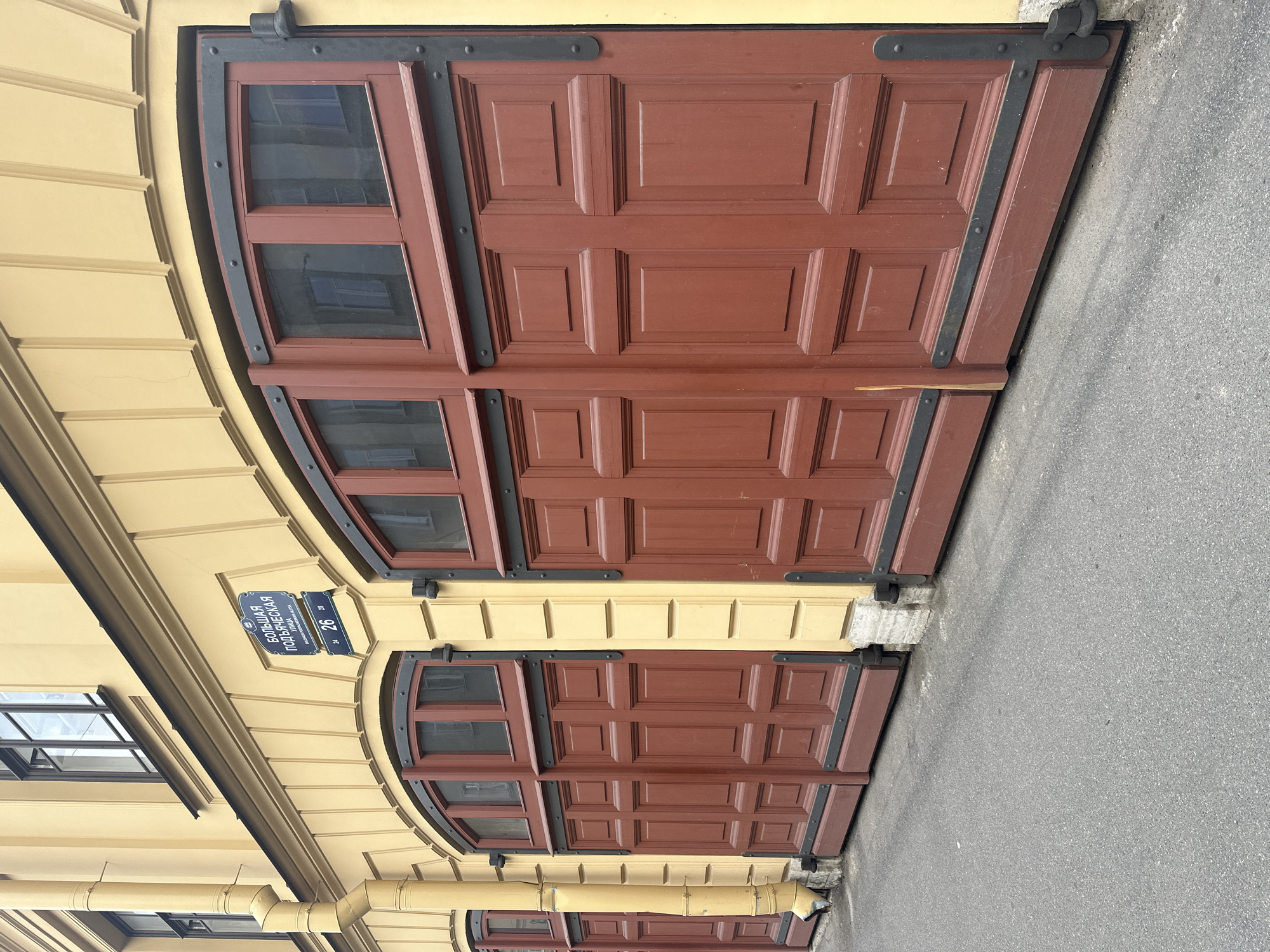 The image captures a section of a building facade in Saint Petersburg, Russia. The building is painted in a light yellow hue and features multiple arched doorways, each fitted with a large, brown, double-door structure. These doors are distinguished by a panel design, segmented by dark-colored hinges or metal strips along the edges. Some of the upper panels on the doors contain window panes. A blue street sign, marked with white Cyrillic text and numbers "24, 26, 28", is affixed above one of the doorways, identifying the location as Больша́я Подъя́ческая улица (Bolshaya Podyacheskaya Street). The ground in front of the building is paved with grey asphalt. The angle of the photograph is canted, with the top of the building appearing to lean to the right.