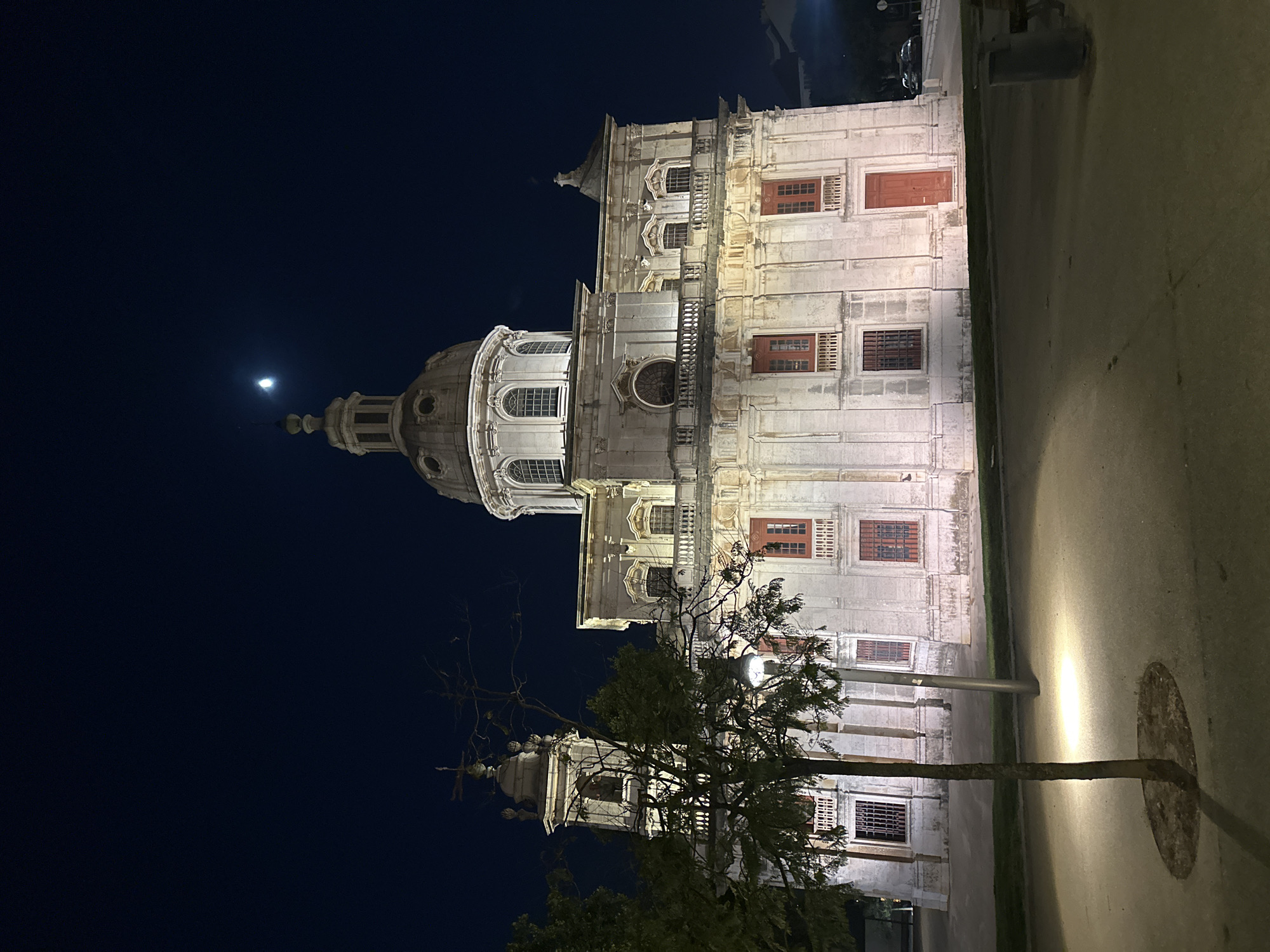 The image depicts a grand building at night, likely a church or a historical monument, bathed in artificial light. The architecture is ornate, with a prominent dome and numerous windows. A tree with sparse foliage stands in front of the building, its branches reaching towards the structure. The sky is dark, indicating nighttime, and a faint moon is visible. The ground in the foreground appears to be a paved area, possibly a street or plaza, with a lamppost casting a bright beam of light. The scene is devoid of people, creating a sense of quiet and solitude. The overall impression is one of historical grandeur and peaceful nighttime ambiance in a European city, possibly Lisbon, Portugal, given the architectural style.