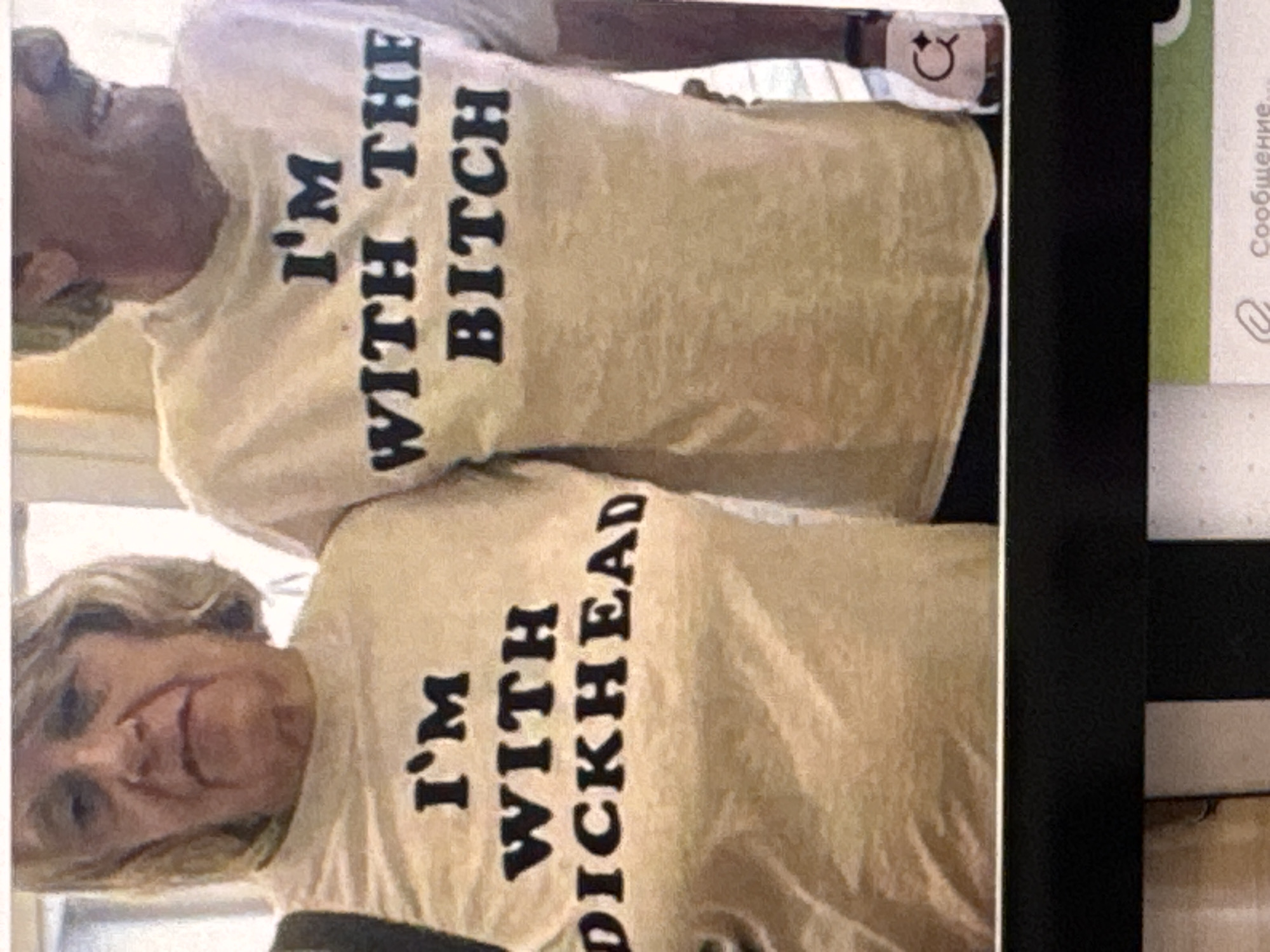 The image shows two people, likely an older couple, wearing matching t-shirts. The t-shirts are light-colored and have black text printed on them. The text on the upper t-shirt reads "I'M WITH THE BITCH". The text on the lower t-shirt reads "I'M WITH DICKHEAD". The people are smiling and appear to be in good spirits. The setting appears to be indoors, possibly a home, with soft lighting. A detail visible on the upper t-shirt is a small, round sticker with a cartoonish face. To the right of the image, a portion of what appears to be a magazine or catalog is visible, with some text in Russian. The overall impression is of a lighthearted and affectionate gesture between the two individuals.