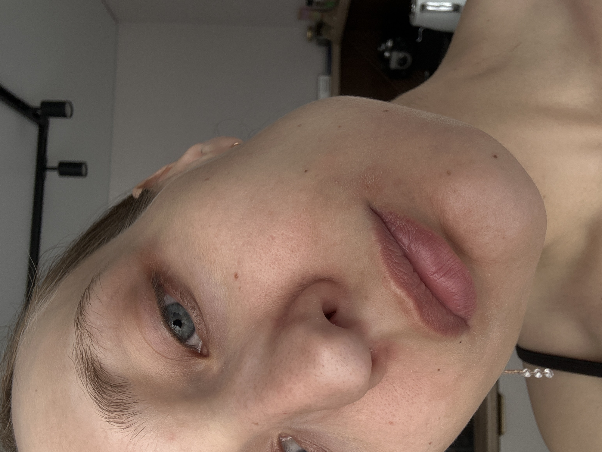 The image is a close-up of a woman's face, rotated to the left. Her skin appears fair, with visible pores and a few small moles. The lips are colored with a pink hue, possibly lipstick. Her eyes are open, and one is clearly visible with a blue iris. The eyebrows are light brown. The background is blurred, but suggests an interior setting with a lamp and a wooden cabinet or door partially visible.
There is no information to place the subject in Подшивалово, Russia.