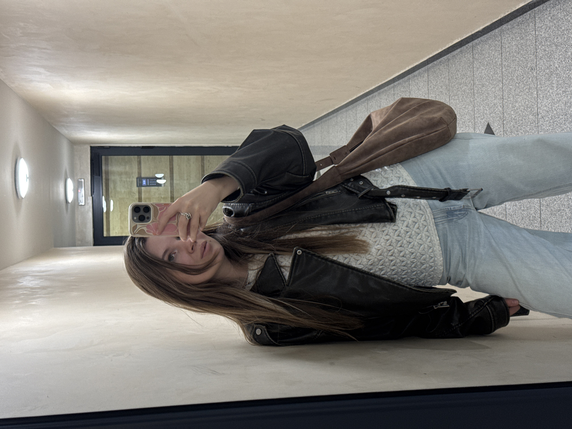 A young woman is taking a mirror selfie in what appears to be an indoor public space. She is wearing a black leather jacket, a light-colored patterned sweater, and light blue jeans. She has a brown shoulder bag slung across her body. She is holding a smartphone up to take the picture. The background consists of a hallway with light-colored walls and tiled flooring, and a doorway is visible in the background. The location is unknown.