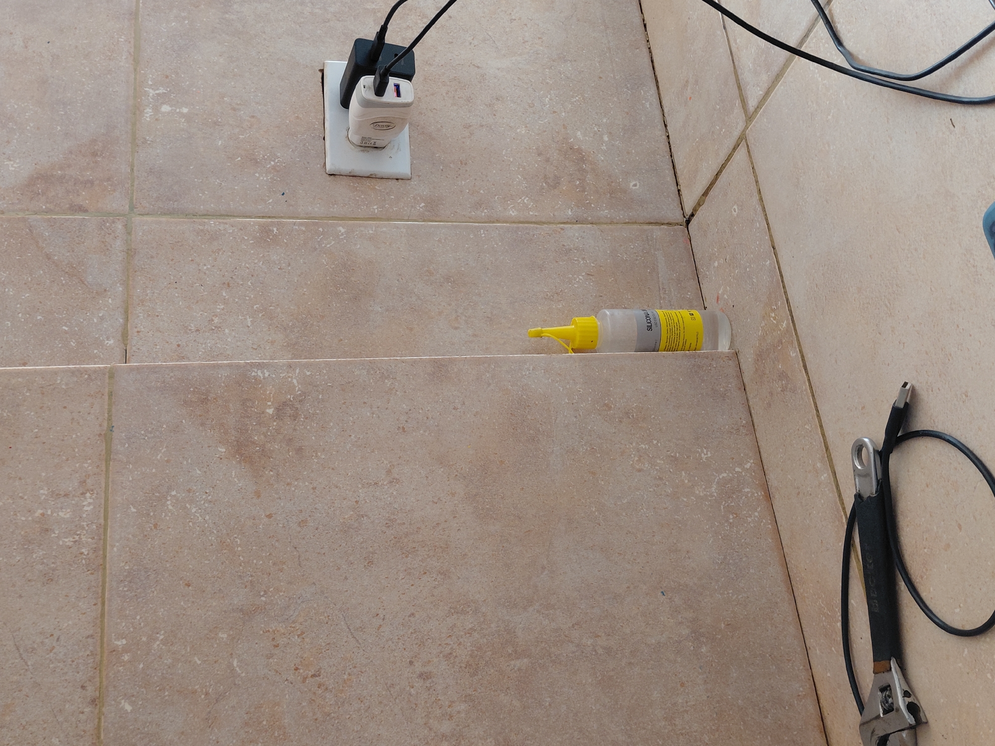 The image captures a static, close-up view of a tiled indoor surface, likely a floor or low wall, in Santa Ana, Venezuela. The tiles are a light, mottled beige-brown color with visible grout lines.
In the upper left corner, an electrical outlet is affixed to the tiled surface. Two electrical chargers, one white and one black, are plugged into the outlet, with their black cables extending upwards and out of frame. The white charger has the brand name "Daway" visible on its side.
A translucent plastic bottle with a yellow pointed cap and nozzle rests horizontally on a lower tiled step or surface in the middle of the frame. Its label is partially visible, clearly showing the word "SILICONA," indicating it contains silicone sealant.
In the lower right part of the image, a black cable is coiled, and a metallic tool resembling an adjustable wrench with a black handle is partially visible. Additional black cables snake across the upper right section of the tiled area. The scene appears to be a utility or work-related space, possibly during or after a repair or installation, given the presence of the tools and sealant. No people or direct activity are depicted.