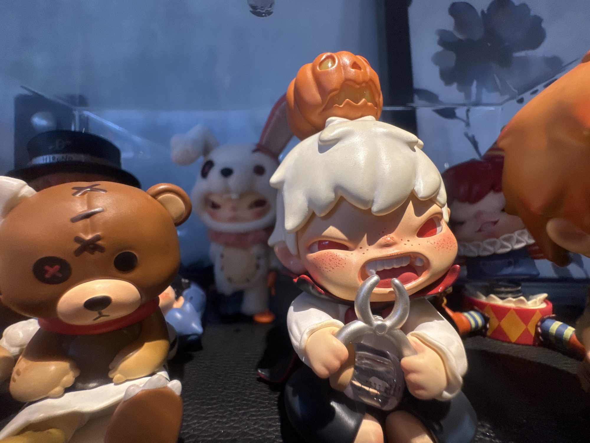 The image shows a close-up of a collection of collectible figurines displayed on a dark surface, possibly a shelf or table. In the foreground, a brown teddy bear figurine with stitched facial features and a red scarf is seated. To its right, a figurine of a young person with white hair, red eyes, and a wide, toothy grin is holding a pair of silver pliers with a small object inside. This figurine is wearing a white shirt and dark pants, with a red cape. Behind these, several other figures are partially visible, including a white bunny-like creature with red accents on its face and a pumpkin-headed figure. The background is a dark, blurred blue, suggesting a shelf or cabinet with lighting. There is no discernible information in the image to suggest a location in Đà Nẵng, Vietnam.
