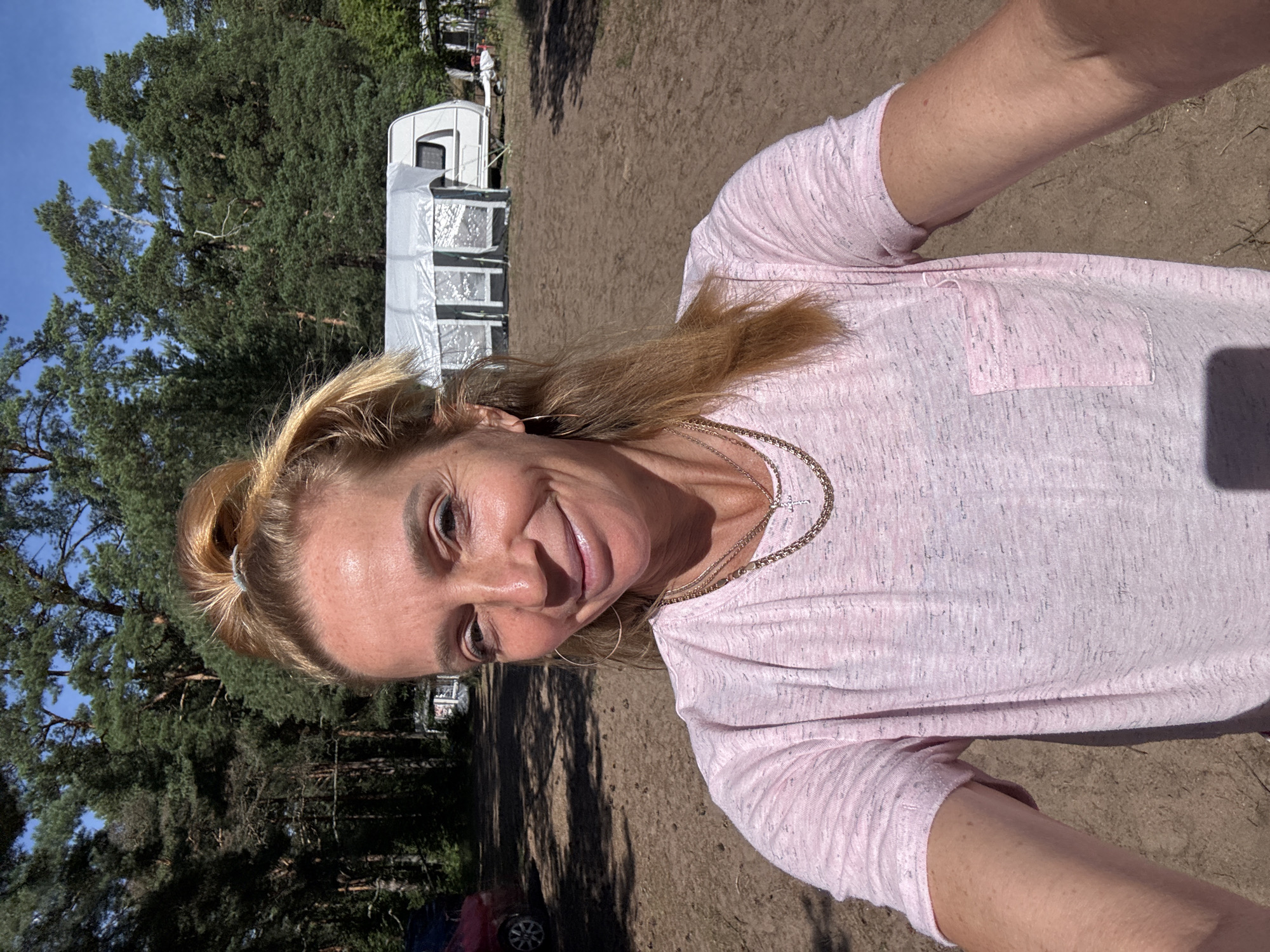 A blonde woman stands in the foreground, taking a selfie outdoors. She has her hair pulled up in a bun and is wearing a pink t-shirt. She is smiling and looking at the camera. In the background, there are trees, a caravan, and a red car. The ground is covered in dirt and sand. The image appears to be taken in a rural or campground setting. There is no indication of the city of Zelenaya Roshcha, Russia in the image.