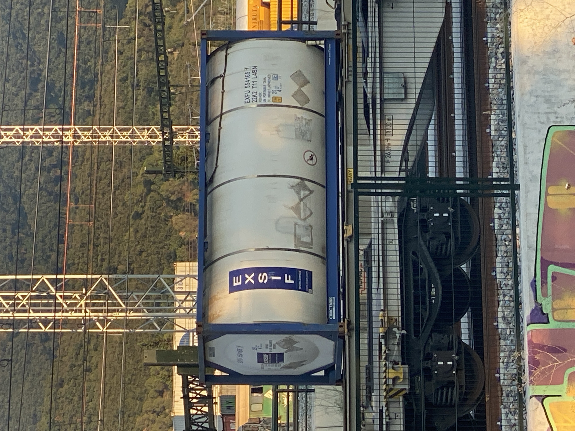 The image shows a train car carrying a large white cylindrical tank container. The container has blue metal framing and blue lettering that reads "EXSIF WORLDWIDE" on one side. On the other side of the container, there is text that includes "EXFU 554165 1 22K2 T11 L4BN". The train car is on a railway track in what appears to be an industrial or transportation hub, with overhead power lines and metal structures in the background. The scene is lit by what seems to be late afternoon or early morning sunlight, casting long shadows. In the foreground, a fence and some graffiti are visible. The image does not contain any people or specific details that would indicate it is located in the city of La Llagosta, Spain.