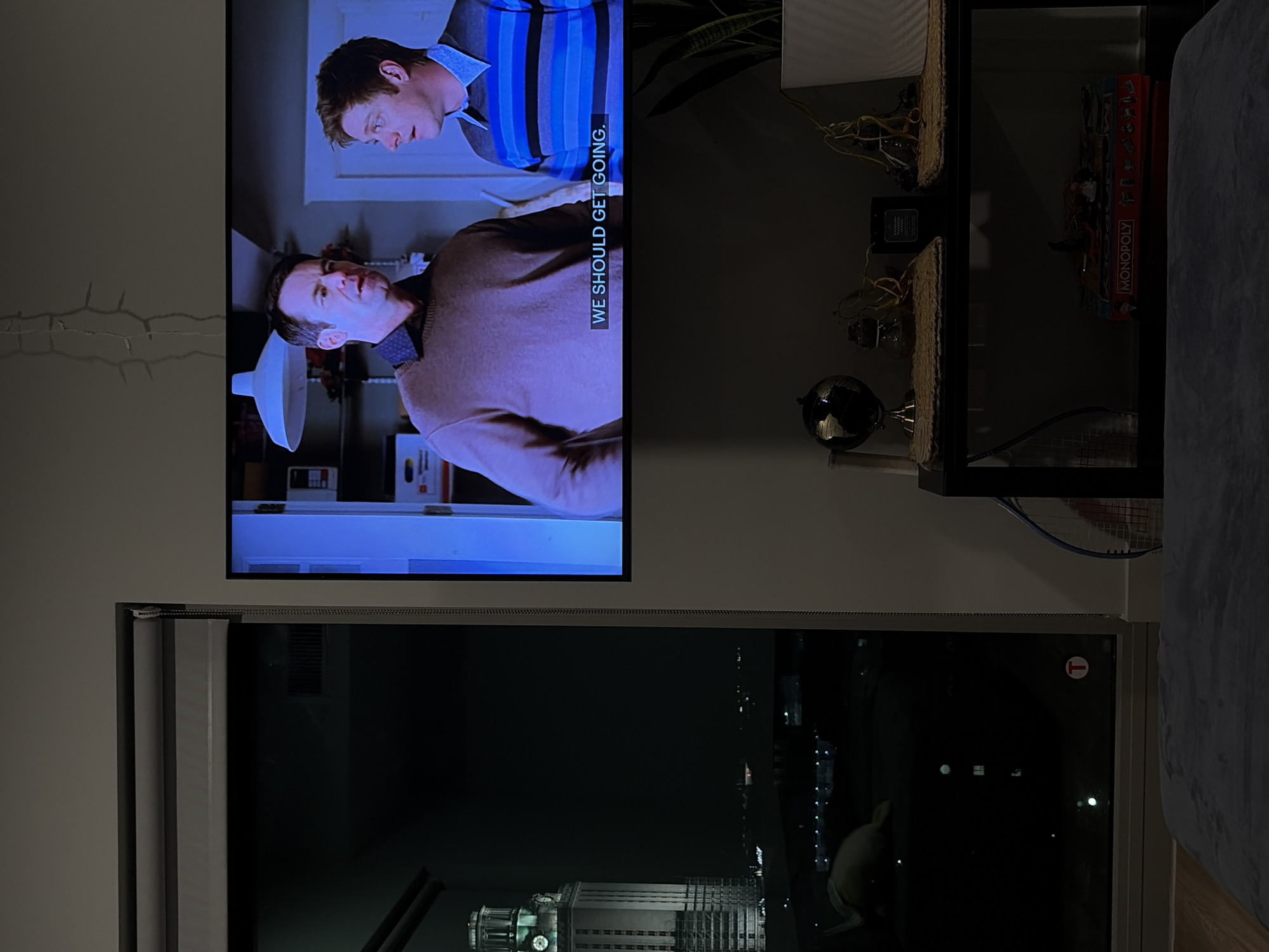A television mounted on a wall is showing a scene with two Caucasian males in a room and superimposed caption that says, "WE SHOULD GET GOING." Below the television is a window showing the night skyline of downtown Austin, Texas, including the clock tower of the Travis County Courthouse. Next to the television is a dark shelving unit containing decorative objects and a Monopoly game box.