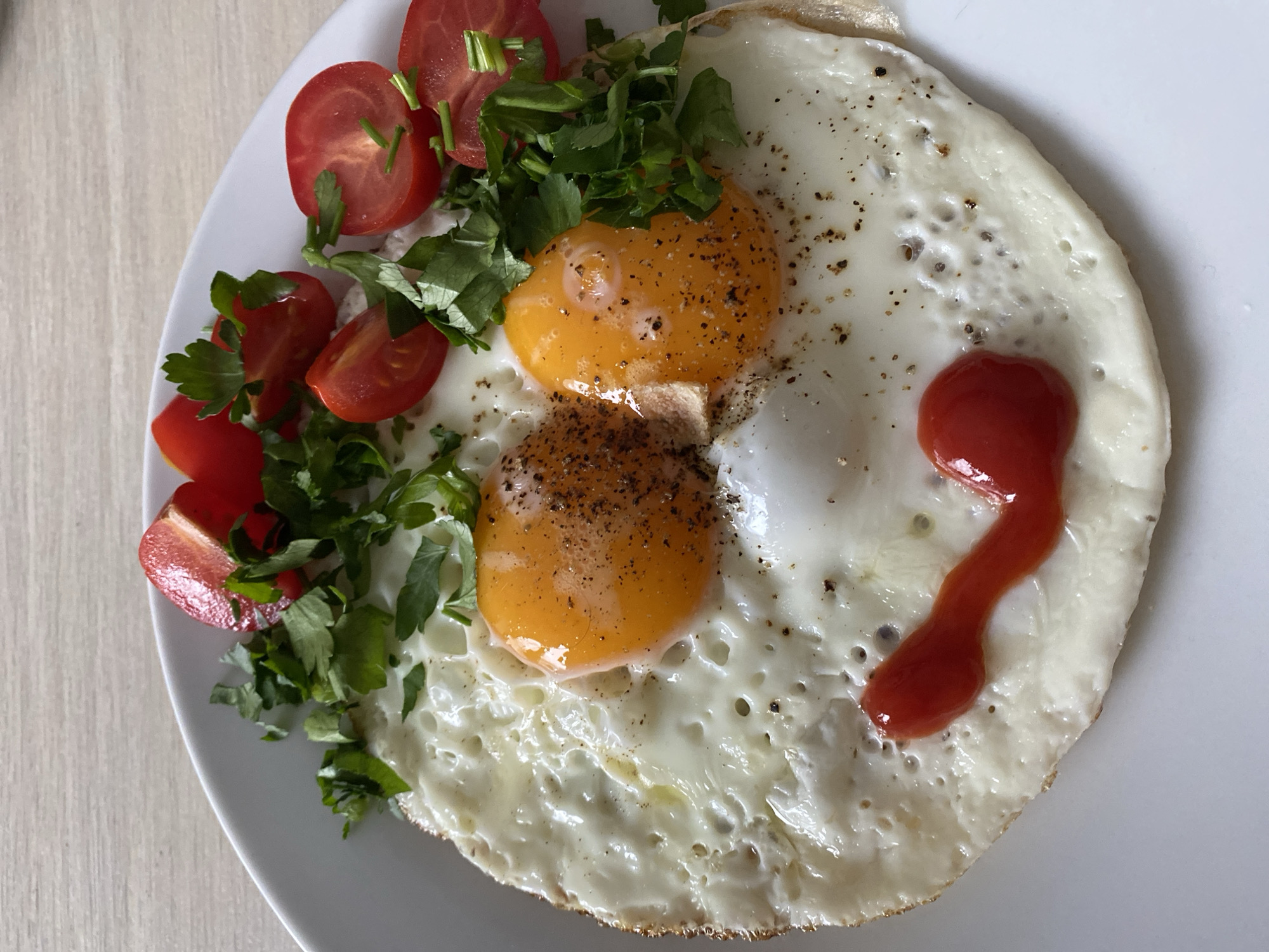 A white plate contains a fried egg, cut cherry tomatoes, and leafy green garnish. The egg is cooked with two yolks, seasoned with pepper, and a squirt of ketchup. The tomatoes are arranged alongside the egg, interspersed with fresh herbs, possibly parsley. The scene is likely indoors on a light wooden table, and there is no verifiable evidence to suggest the location is specifically in Chaikovsky, Russia.